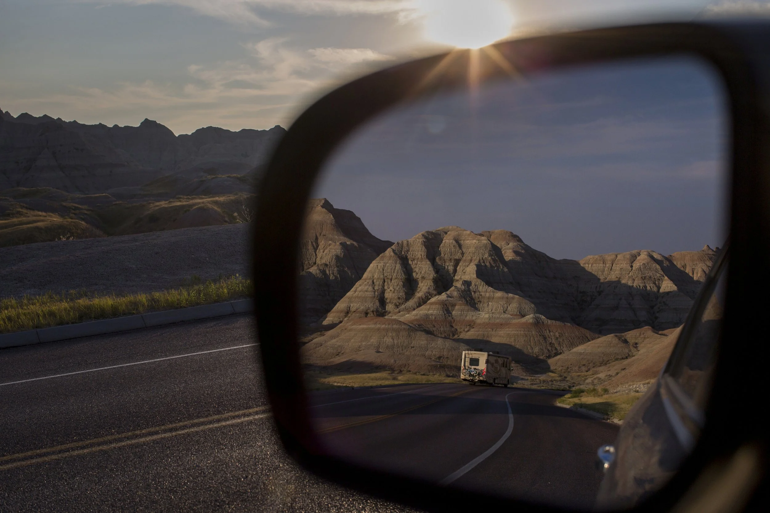 View of the Badlands National Park through a car's side mirror with the sun setting in the background.