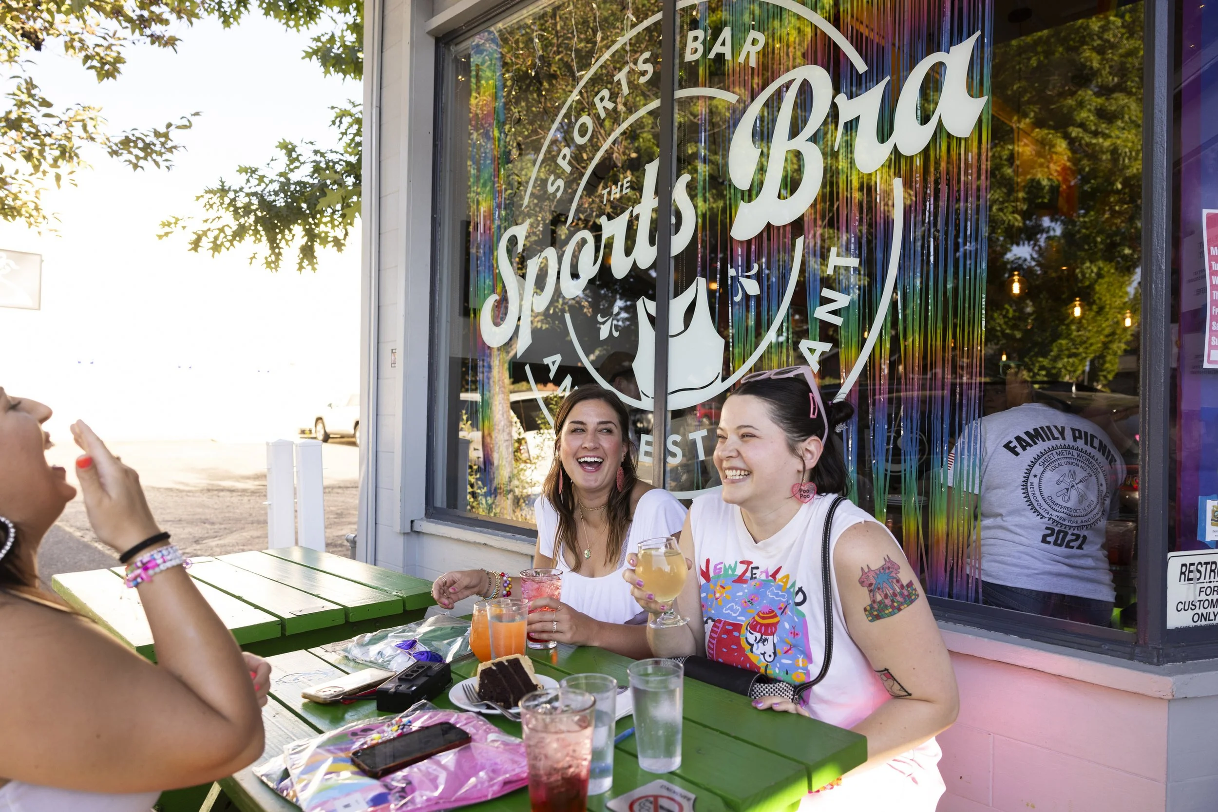 Two smiling women sitting at a green picnic table outside The Sports Bra bar and restaurant, with drinks and a slice of chocolate cake on the table, during the daytime in Portland, Oregon.