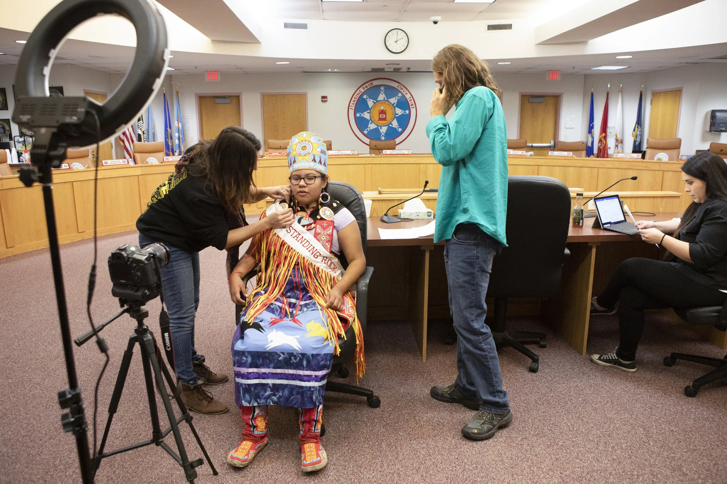 A Native American woman in a conference room. Several people, including one talking on a cellphone and another working on a laptop, are present. The room has a long table, chairs, and flags in the background.