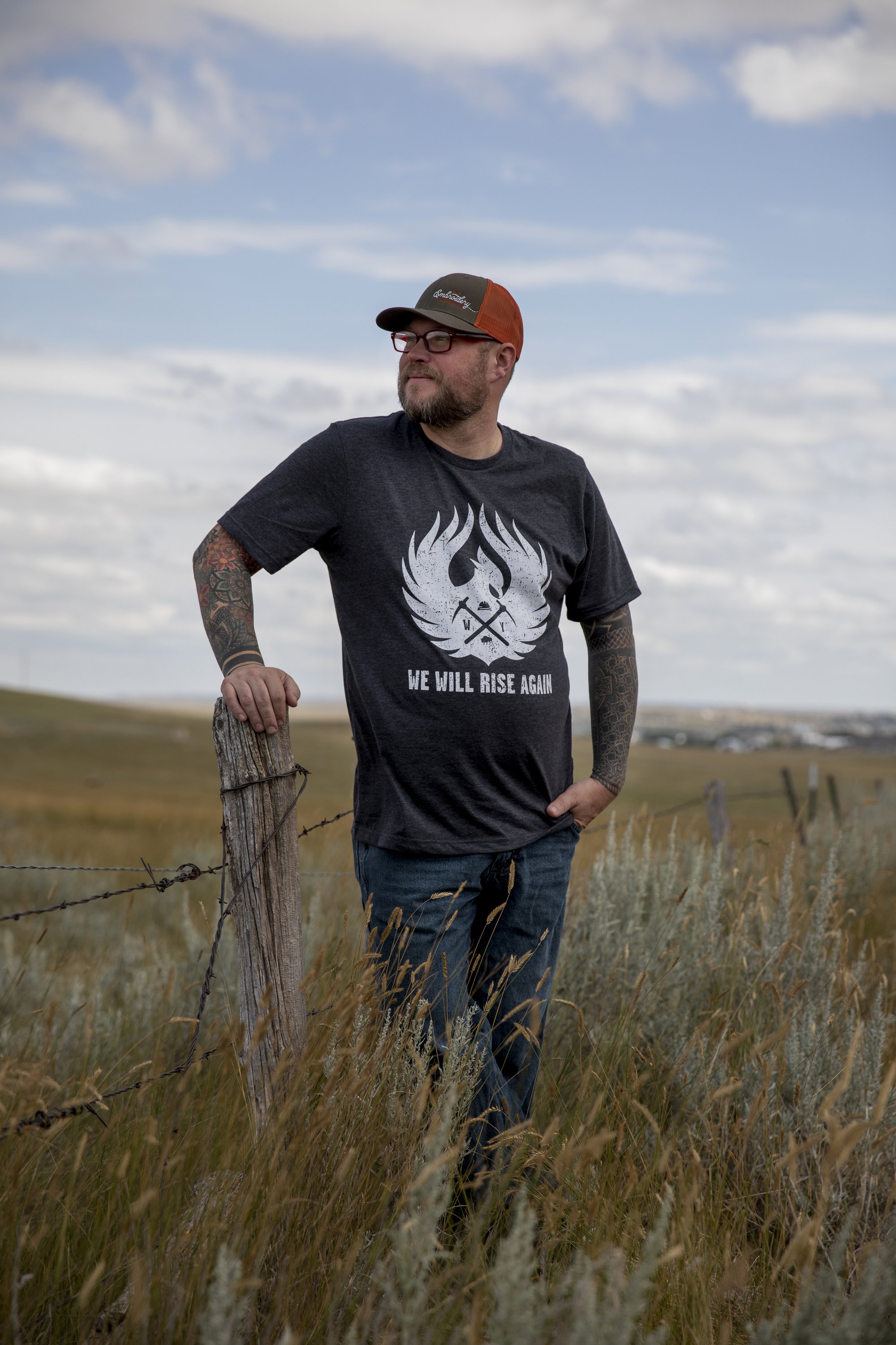 A man with tattoos on his arms and beard standing outdoors in a grassy field, leaning on a wooden fence post, wearing a black T-shirt with a white graphic and the words "We Will Rise Again," a trucker hat, and jeans, with a cloudy sky in the backgrou
