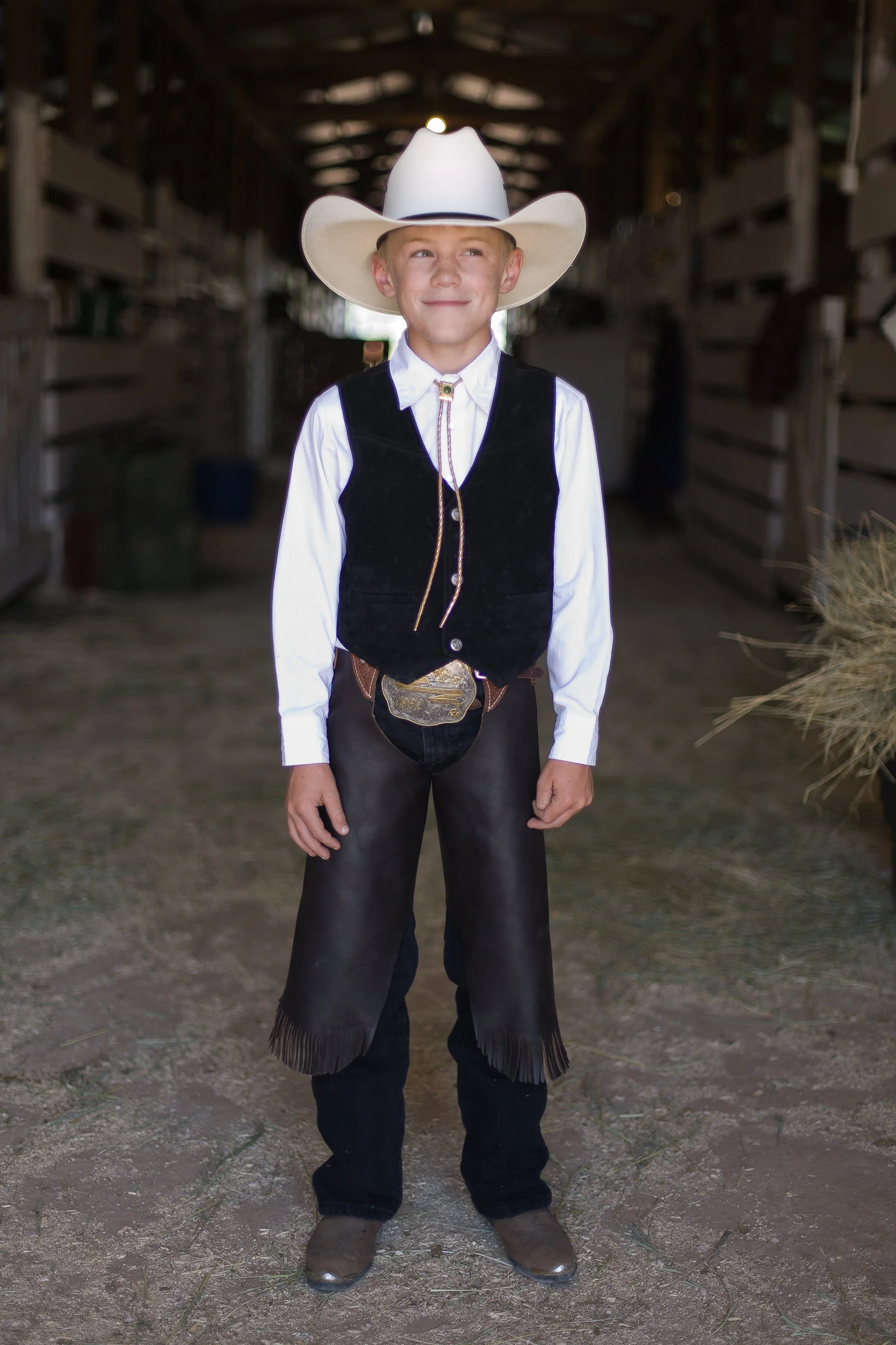 Young boy dressed in cowboy attire, standing inside a barn, wearing a white cowboy hat, white shirt, black vest, and leather chaps.