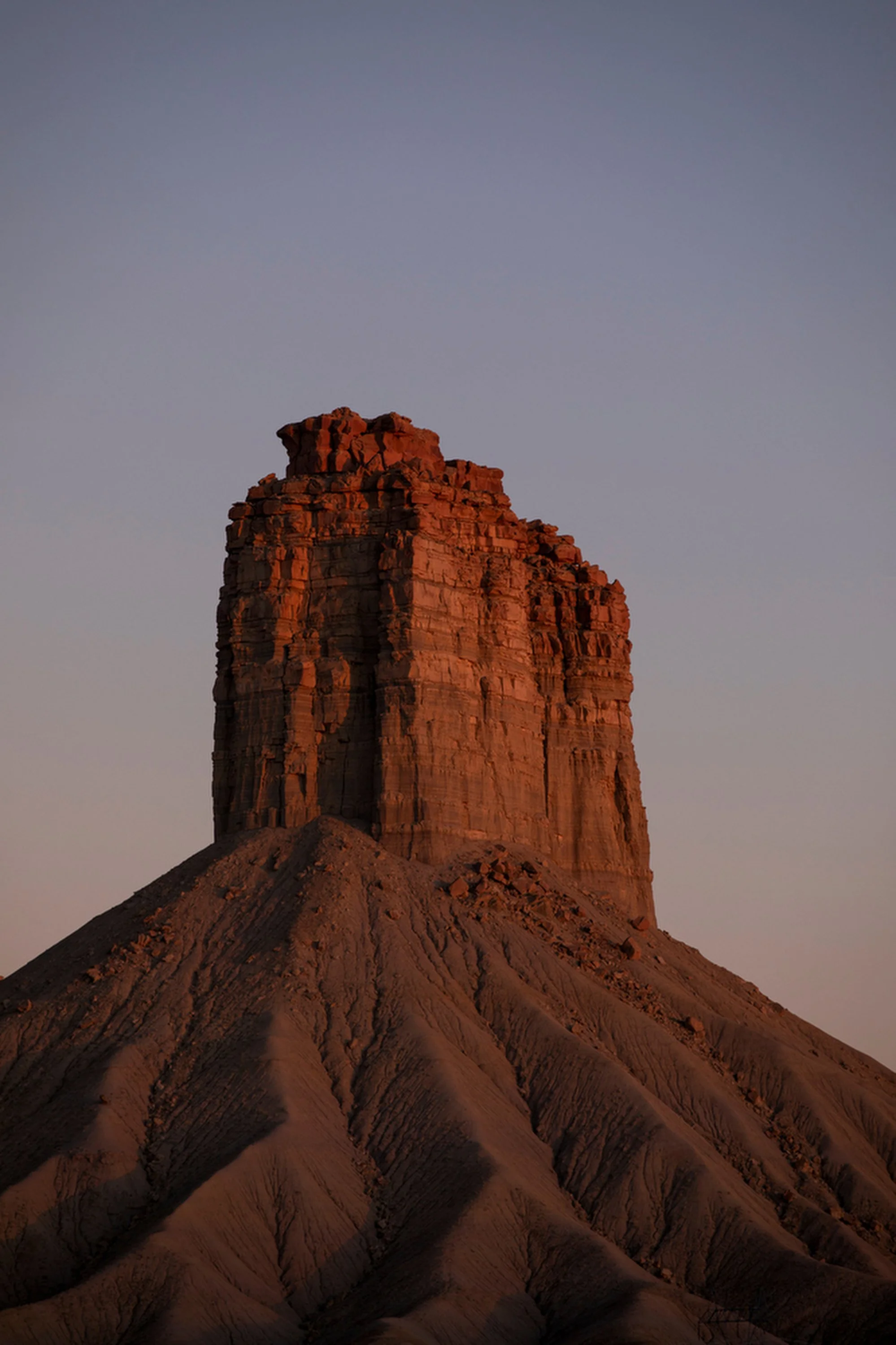 A tall, rocky mesa with a flat top, illuminated by the setting or rising sun, with rugged terrain at its base and a clear sky in the background.