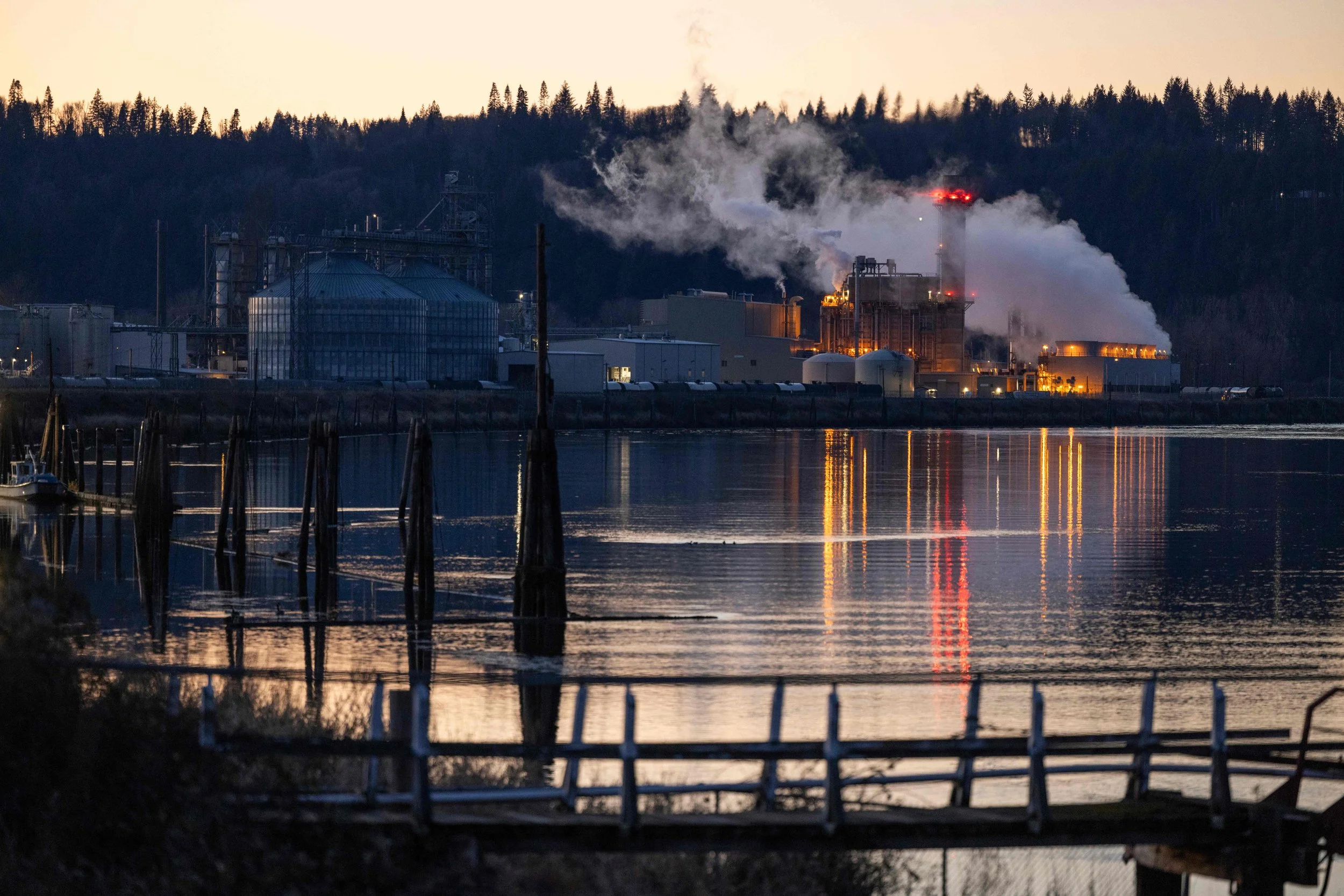 Industrial plant near a body of water with smoke and steam emissions during the evening, with a forested hillside in the background.