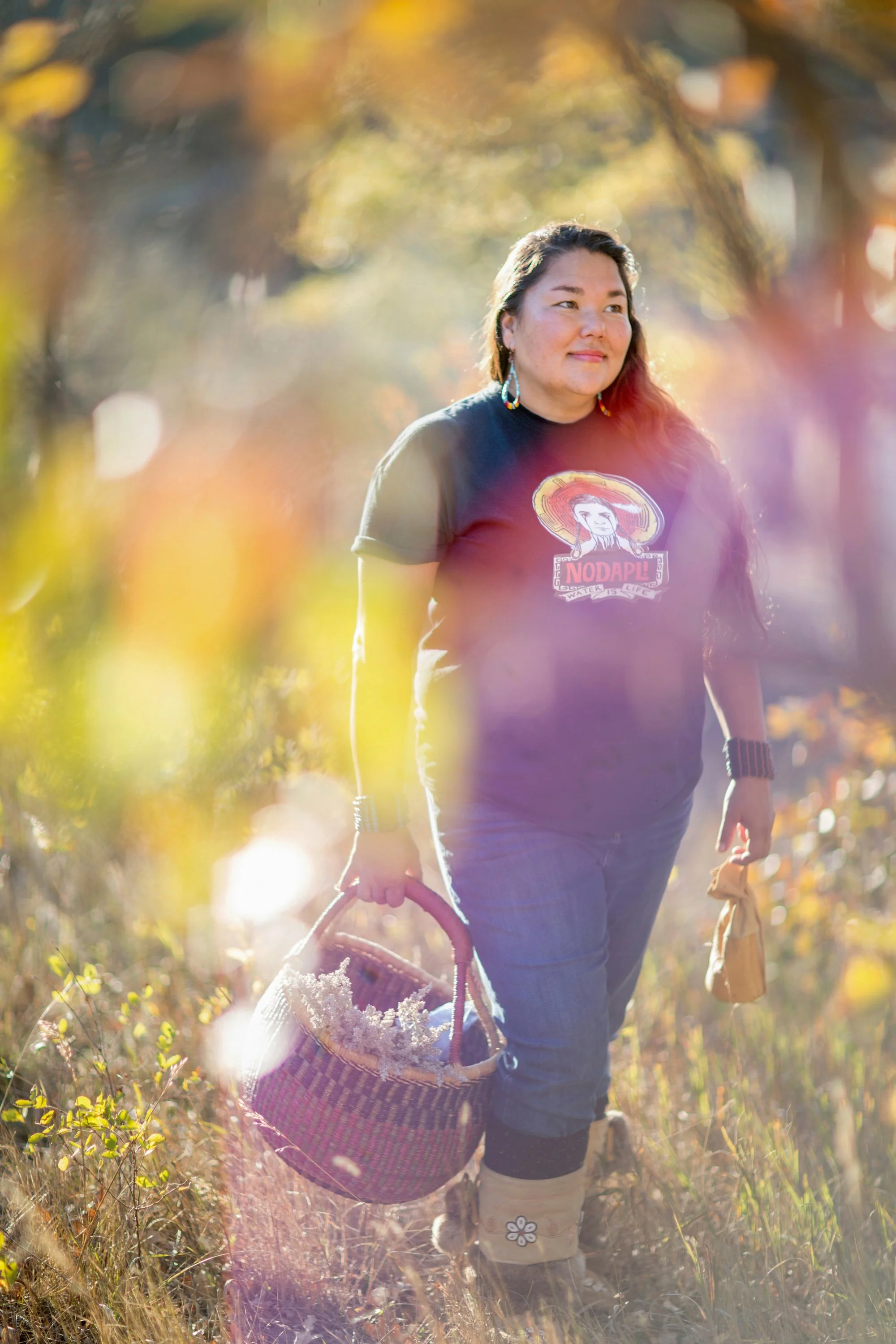 A woman walking in a fall outdoor setting holding a basket and a bag, wearing a black t-shirt with a mariachi image and the word 'NODAPL', jeans, and beige boots, with sunlight filtering through trees in the background.