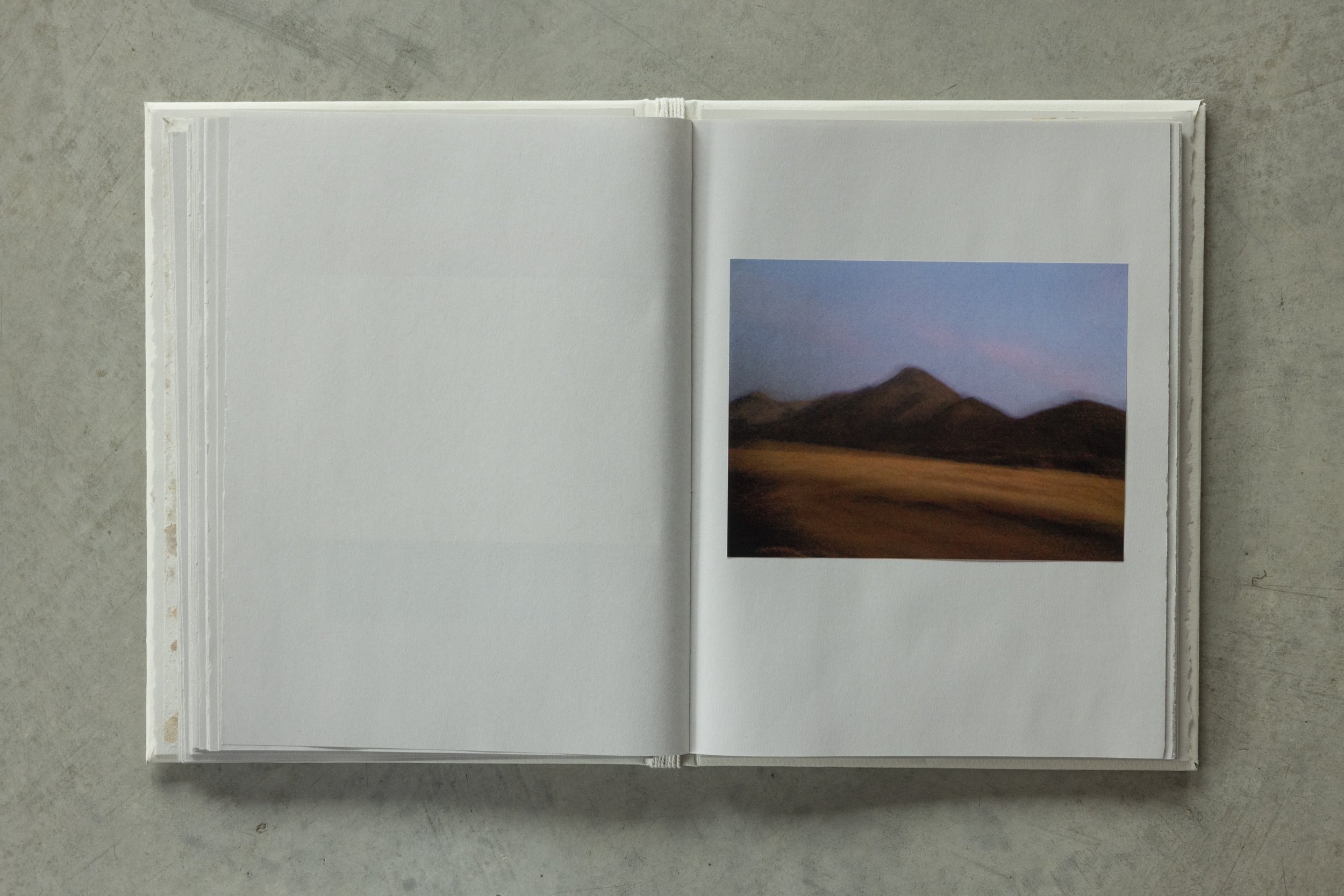 Open white photo album lying open on a gray surface, showing a landscape photograph of mountains and a colorful sky on the right page.