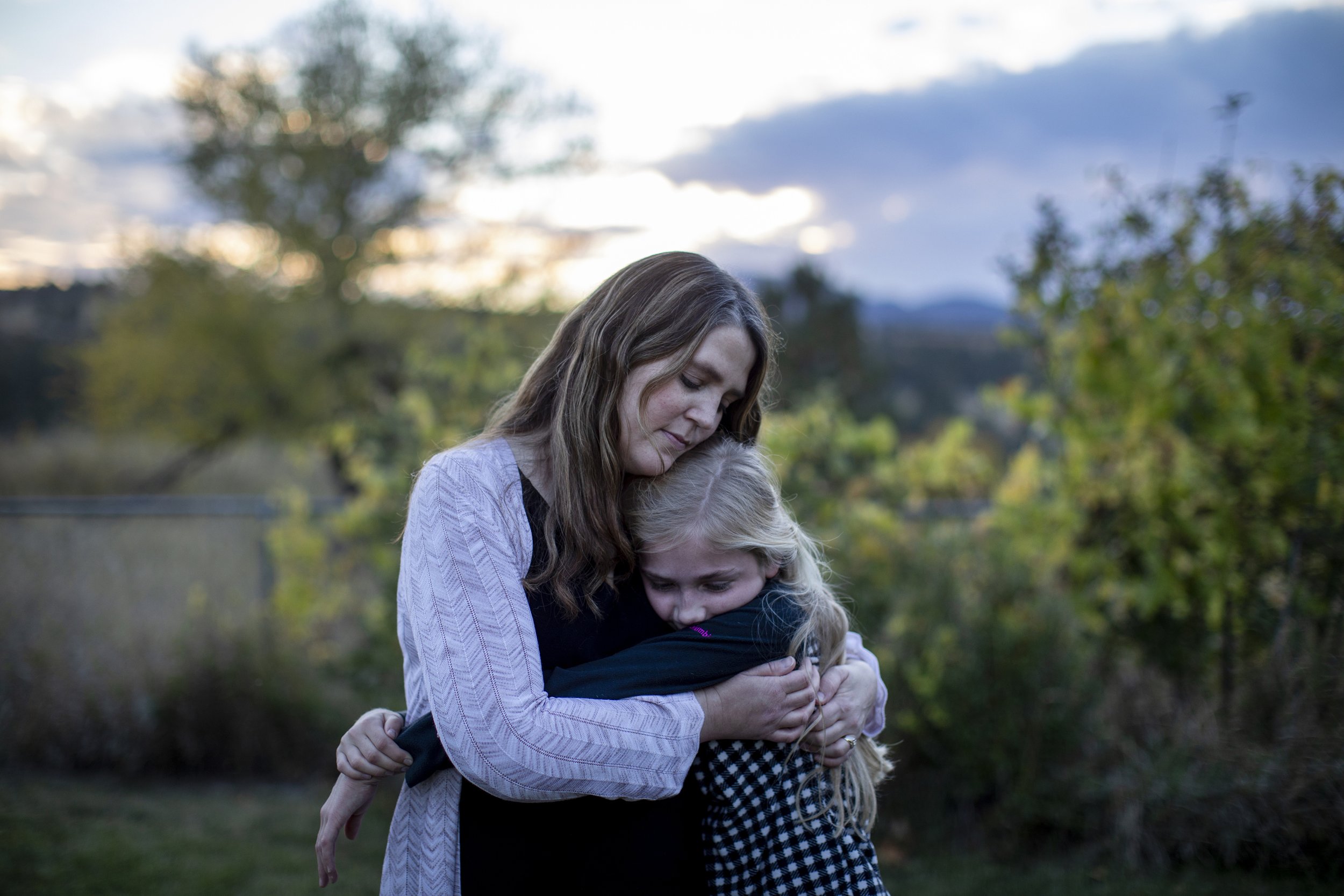A woman hugging a young girl outdoors during sunset with trees and cloudy sky in the background.
