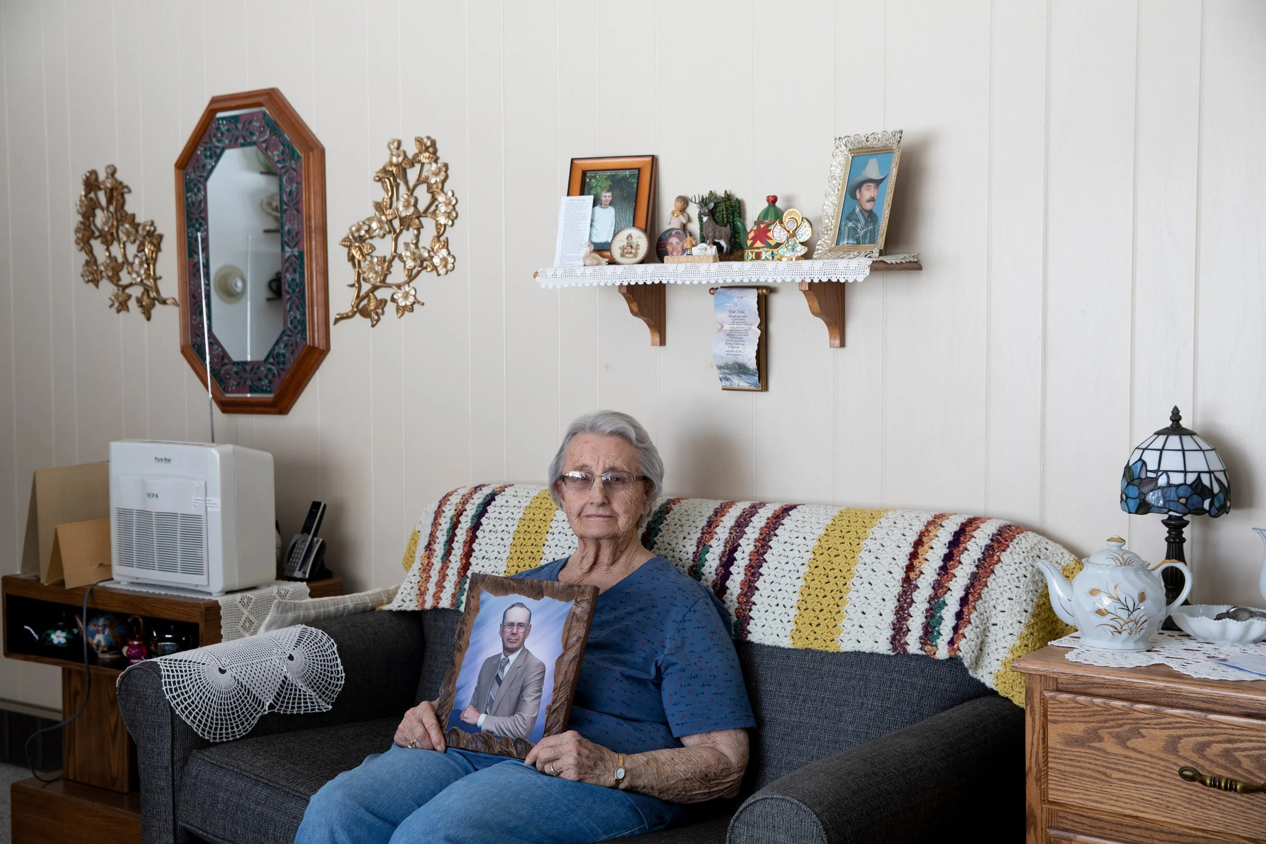 An elderly woman sitting on a couch holding a framed photograph. The room has a white wall with shelves displaying framed photos, decorative figurines, and a small picture. A lamp with stained glass shade and a teapot are on a side table in Mobridge.