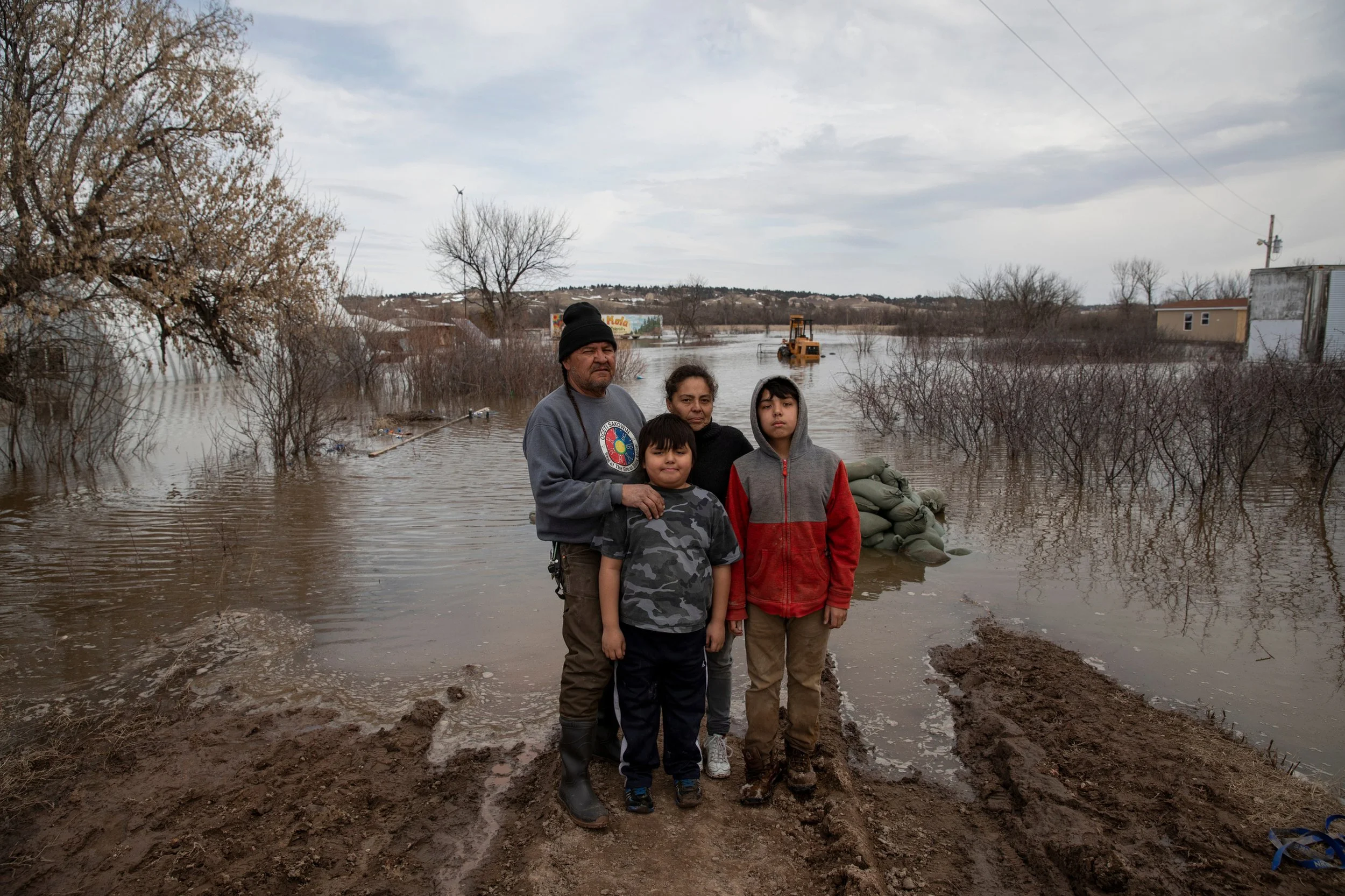 A family standing on muddy ground in front of floodwaters, with trees, a tractor, and buildings in the background, under an overcast sky in South Dakota.