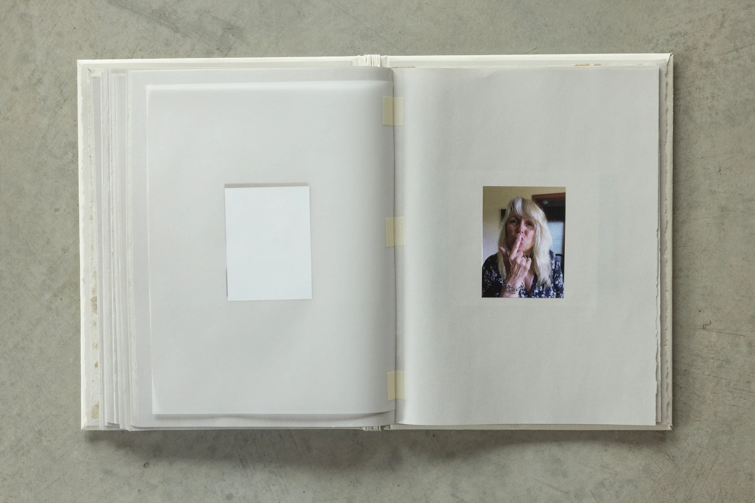 Open scrapbook or photo album with blank pages and a small photograph of a woman with blonde hair, making a shushing gesture, taped onto the bottom right corner of the right page.