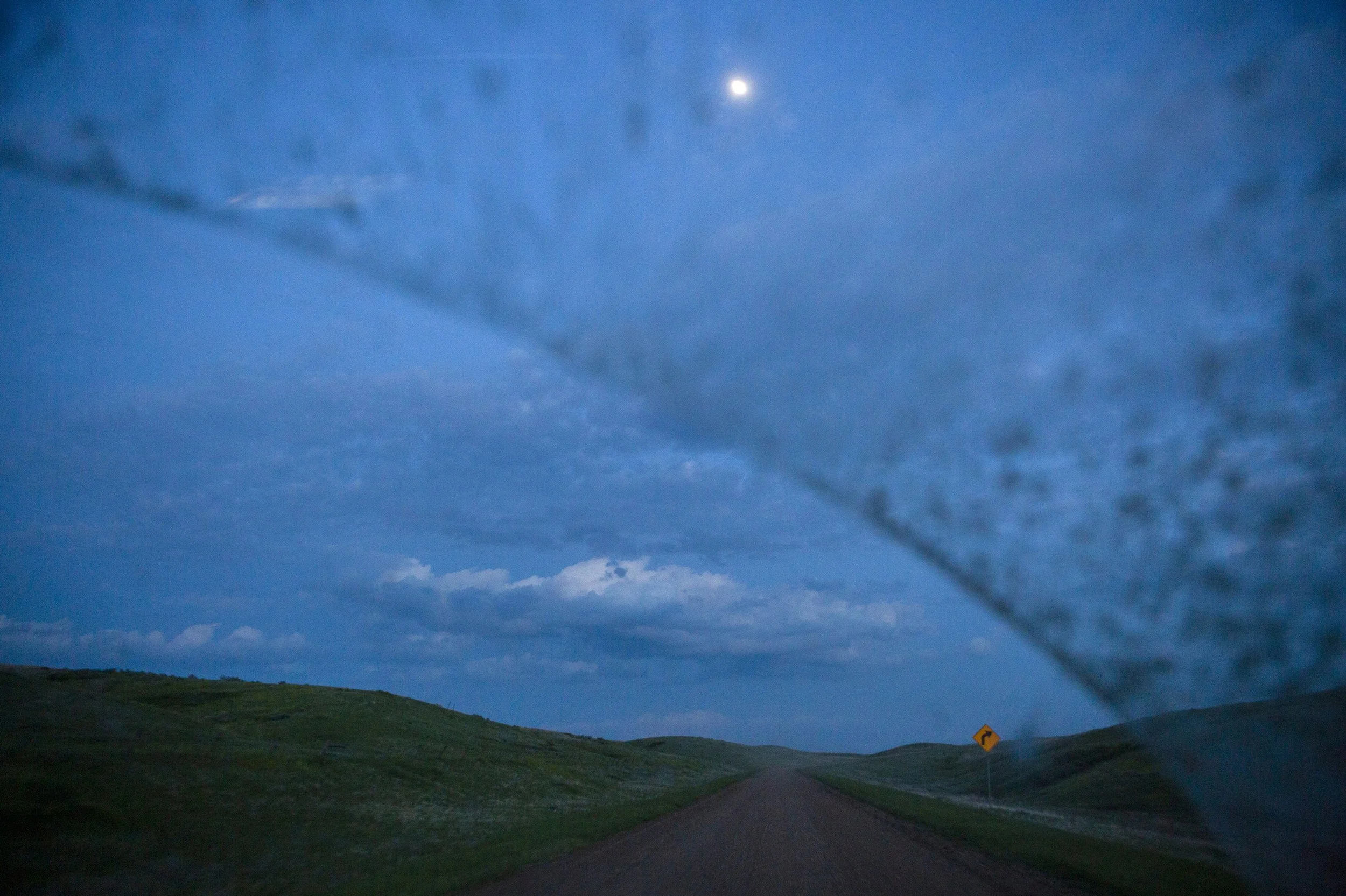 A winding road in a rural area under a blue and cloudy sky at dusk, with a sign indicating a right turn.