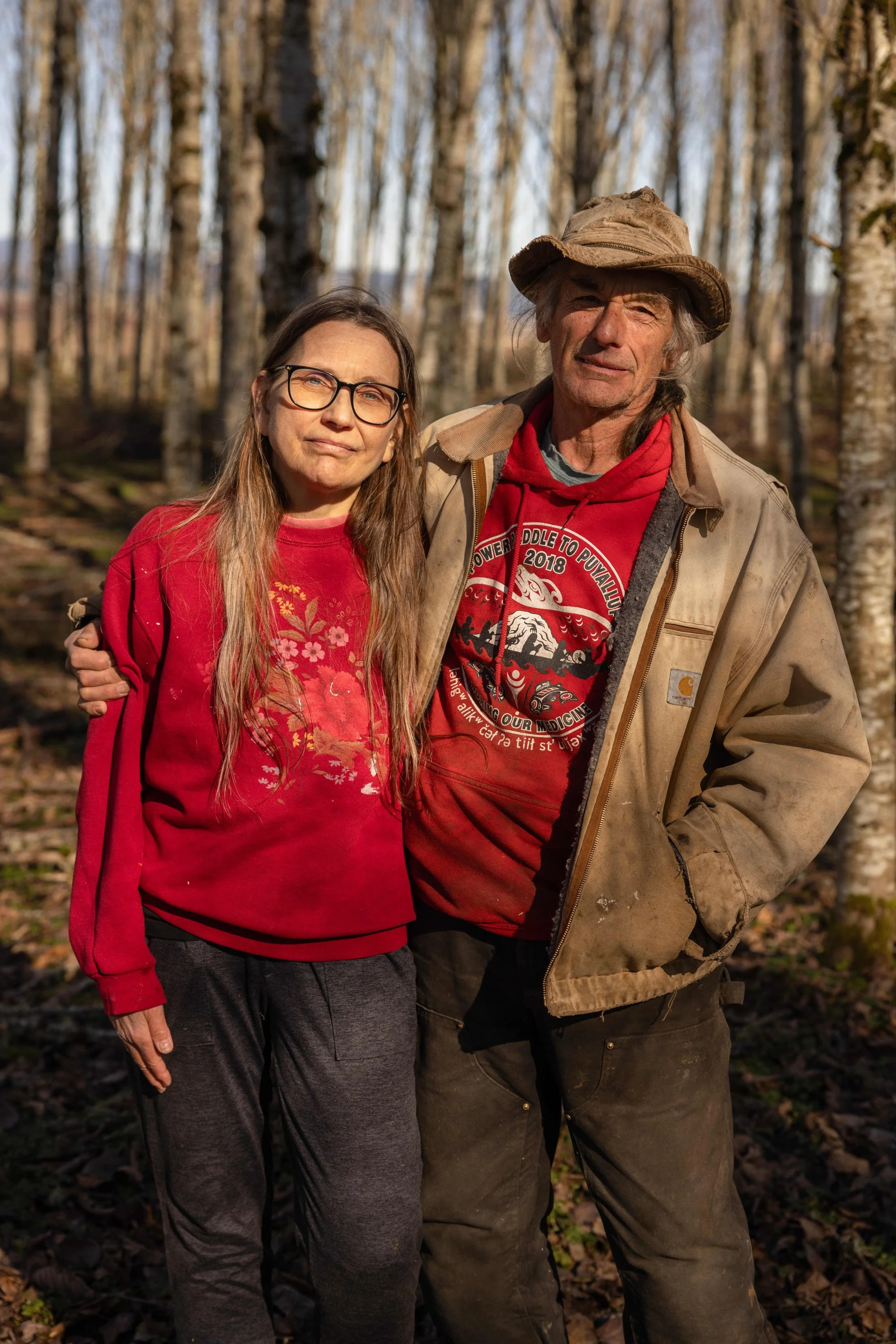A woman and a man standing outdoors in a forest with tall trees in the background, wearing casual clothing, and posing for the photo.