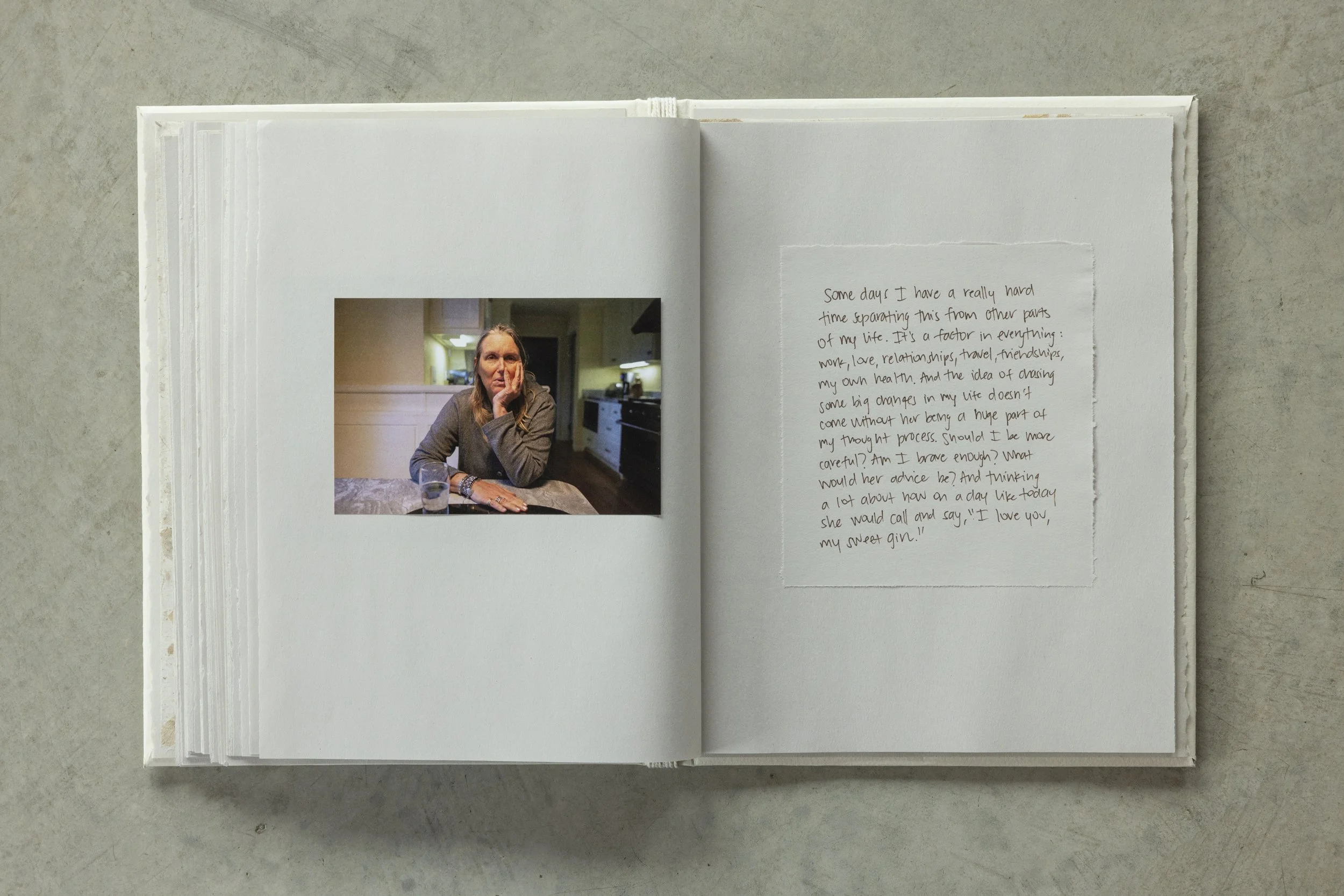 Open scrapbook displaying a photograph of a woman sitting at a kitchen table, resting her chin on her hand, with a handwritten note on the adjacent page.