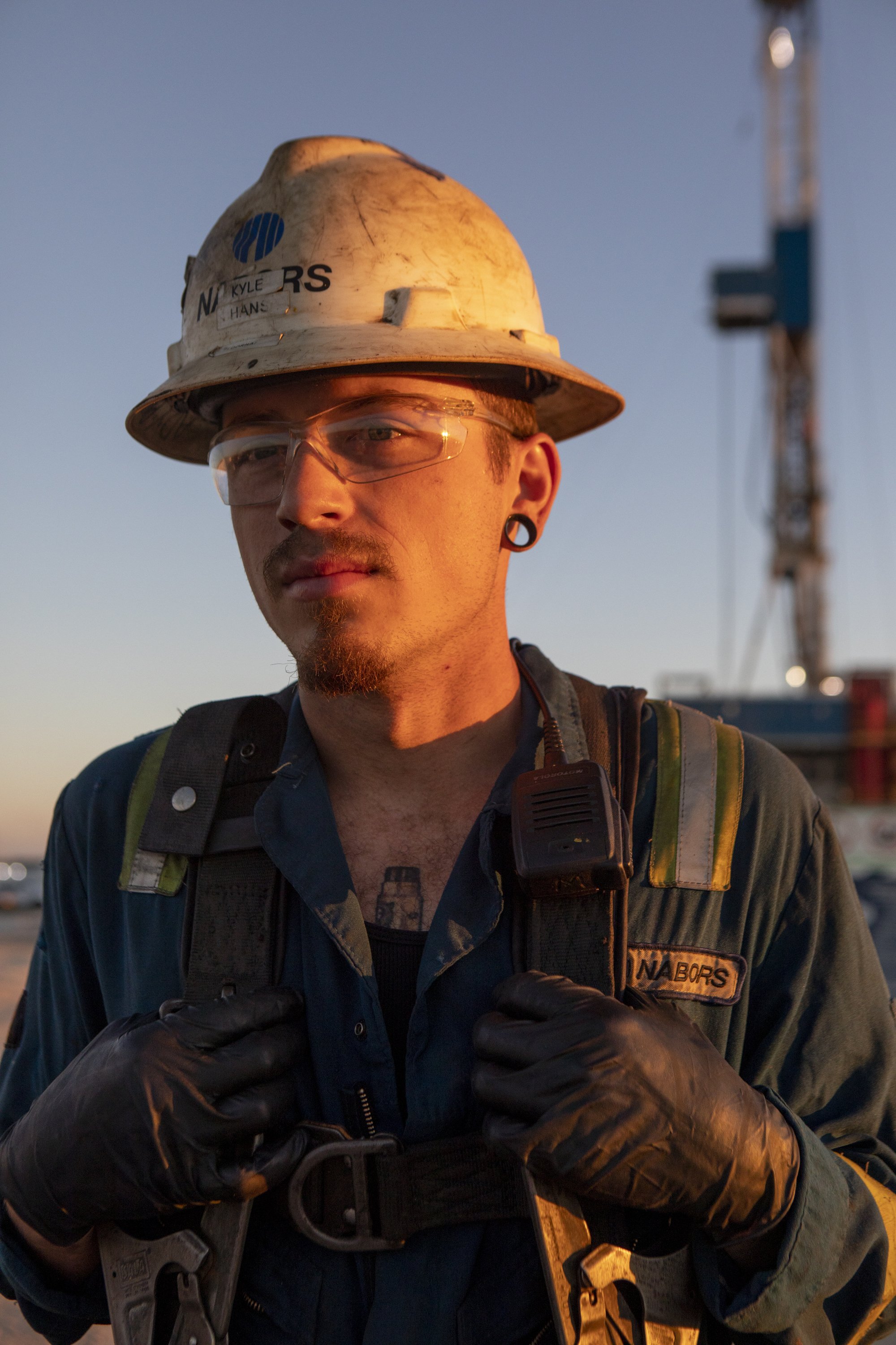 A male worker wearing a white hard hat, safety glasses, and gloves, with a ladder tower in the background during sunset in Wyoming.