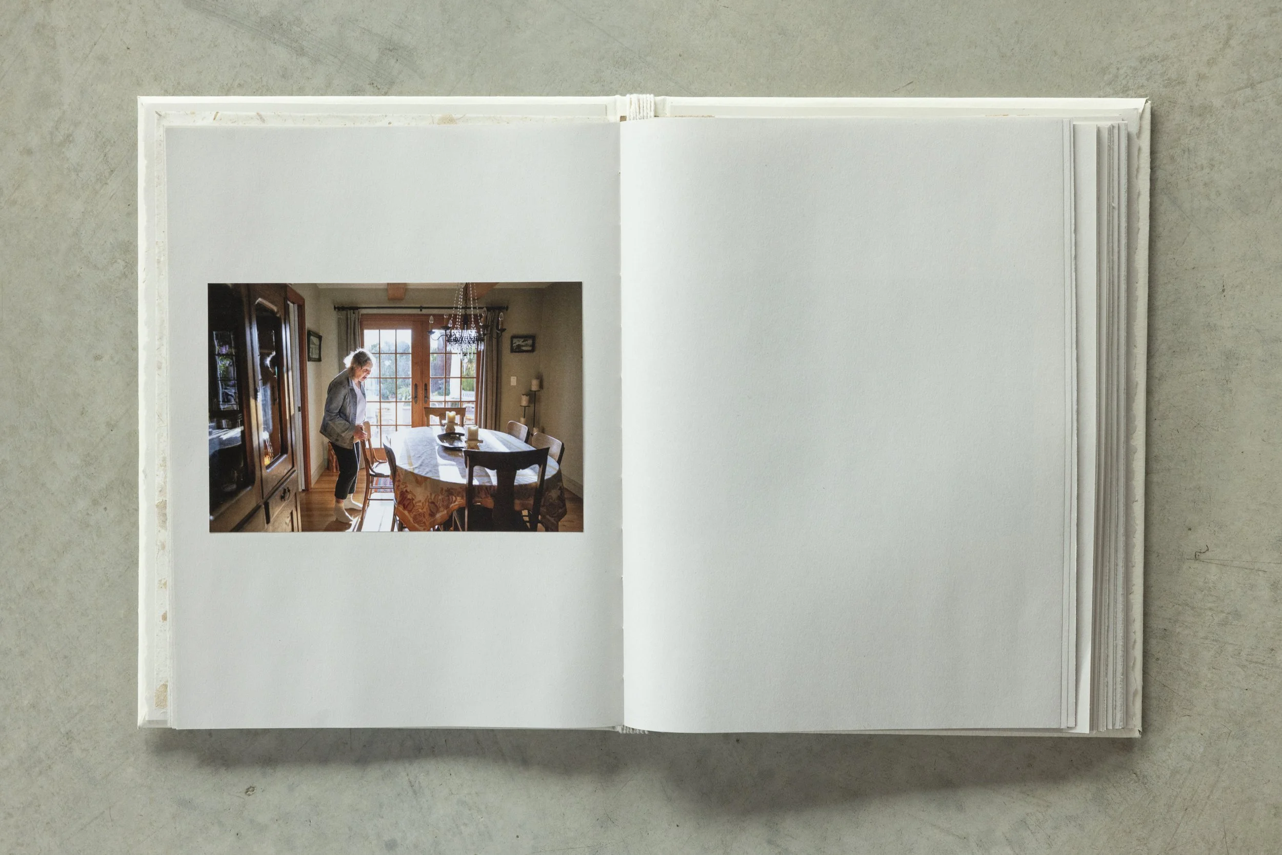 A photo book open to a page with a photo of a woman in a dining room with a large window, wooden furniture, and chandeliers.