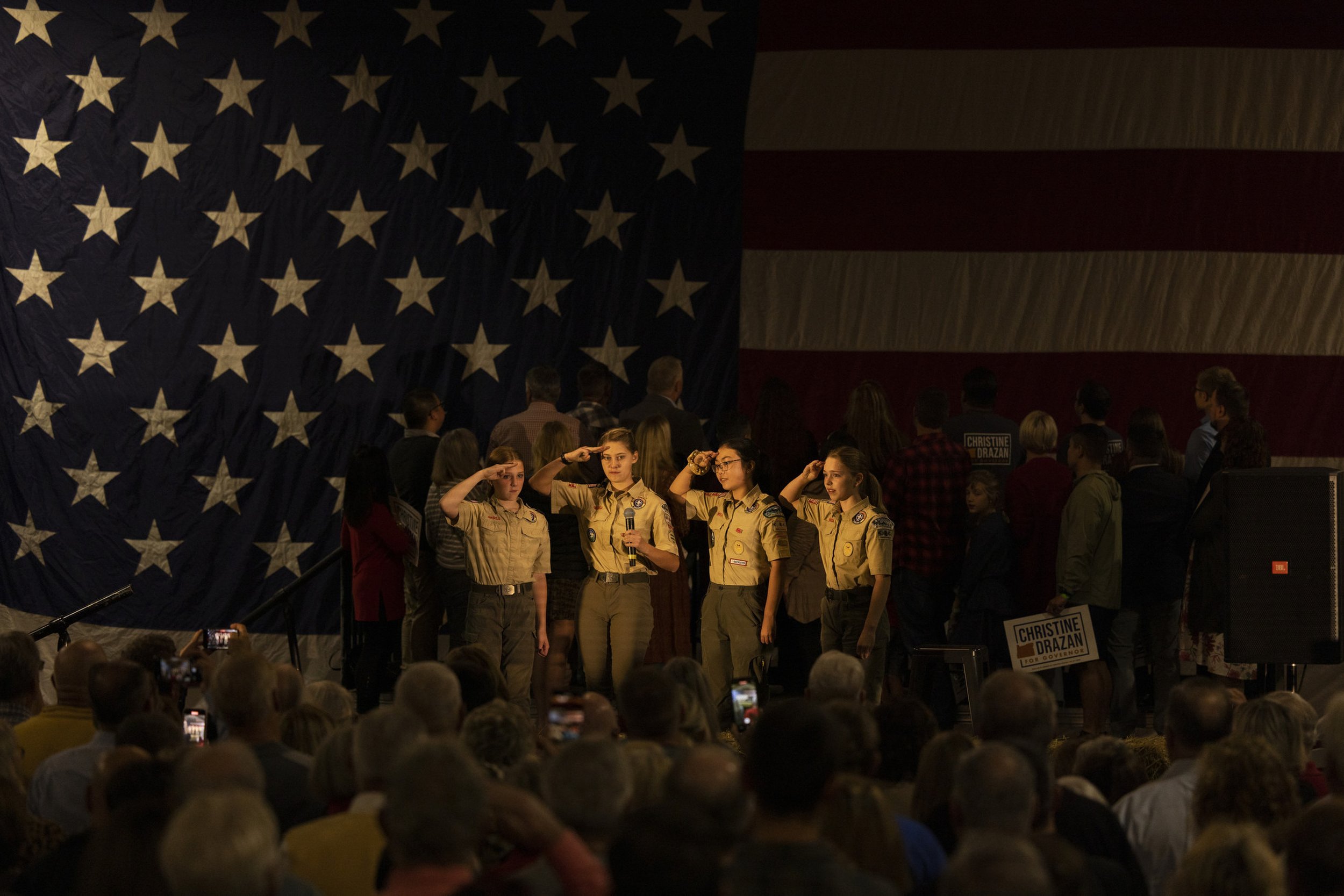 A large American flag hangs in the background of a crowd at a political event. In front, four young girls in matching Girl Scout uniforms are saluting as they stand in a row. Several people in the audience are watching and taking photos.