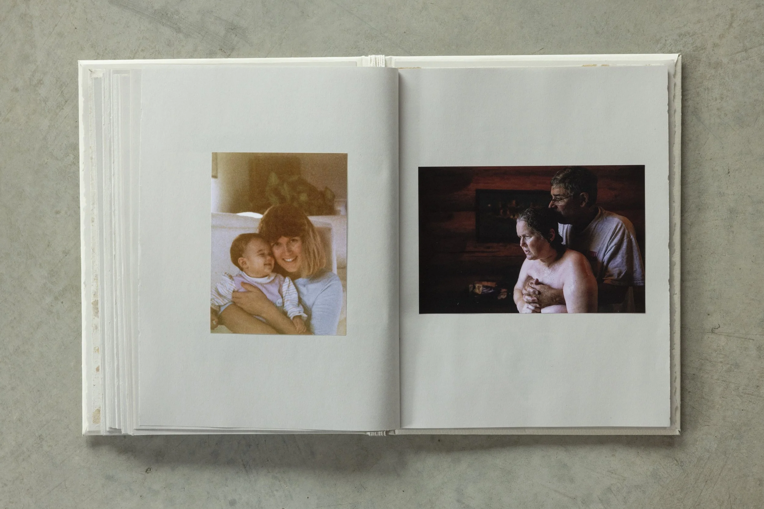 Open photo album showing two pages with printed photographs on a light grey surface. The left page features a picture of a woman with shoulder-length hair holding a smiling young boy who is looking at her. The right page shows a woman with short dark