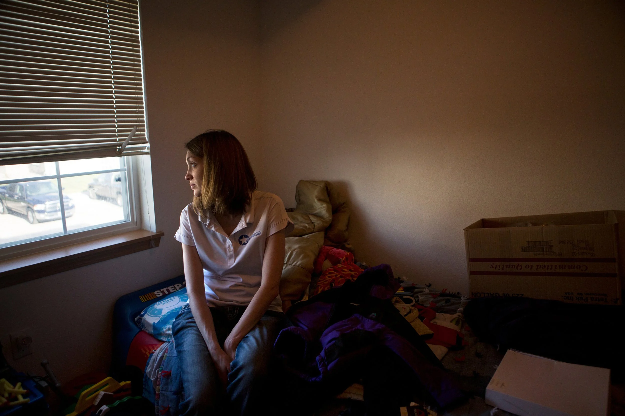 A young woman with shoulder-length brown hair, wearing a white polo shirt, sitting on a cluttered bed in a dimly lit room near a window with blinds. Various objects are scattered around.