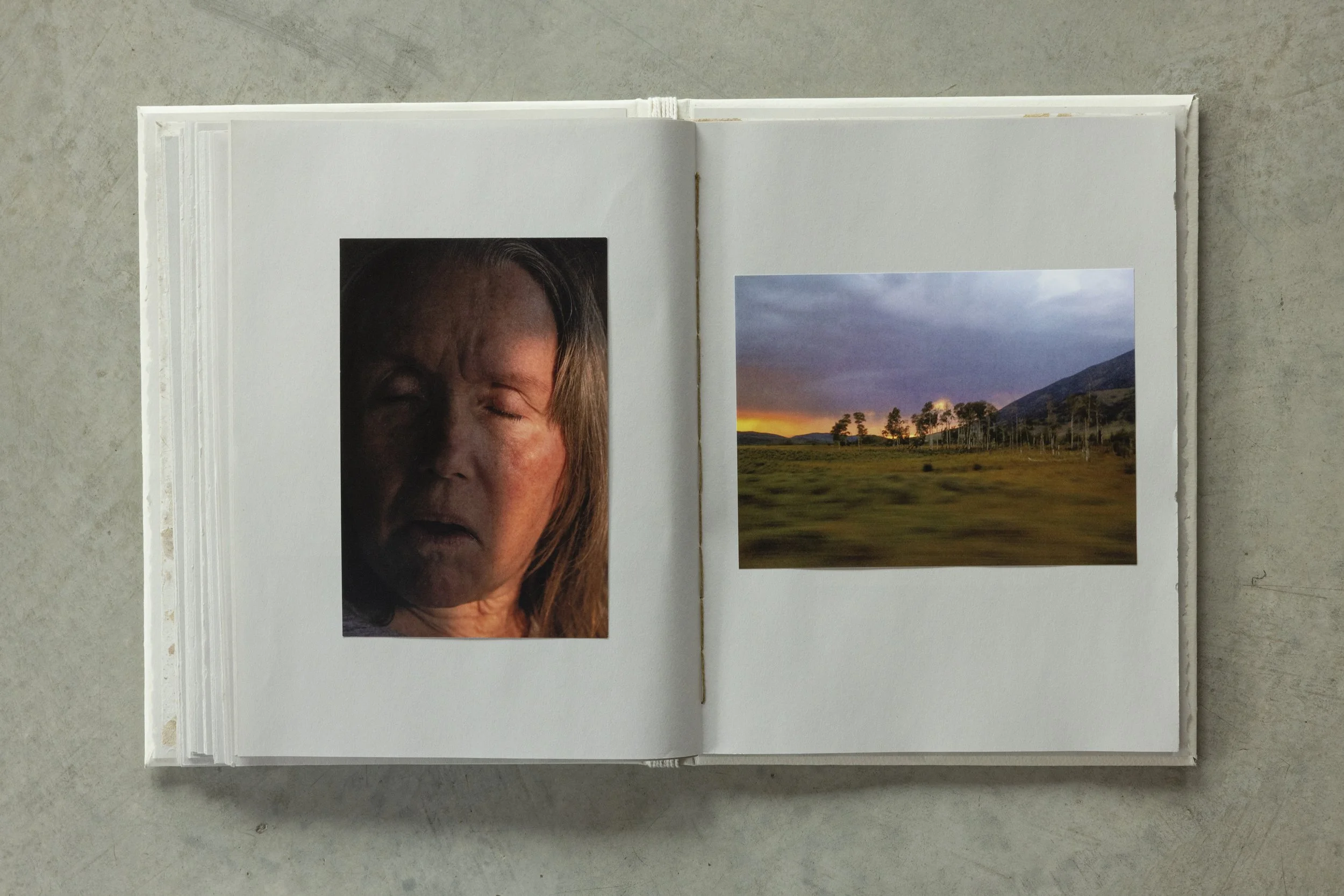 Open photo album with a large portrait of a woman with closed eyes on the left page and a landscape photograph of a sunset over trees and mountains on the right page, placed on a gray surface.