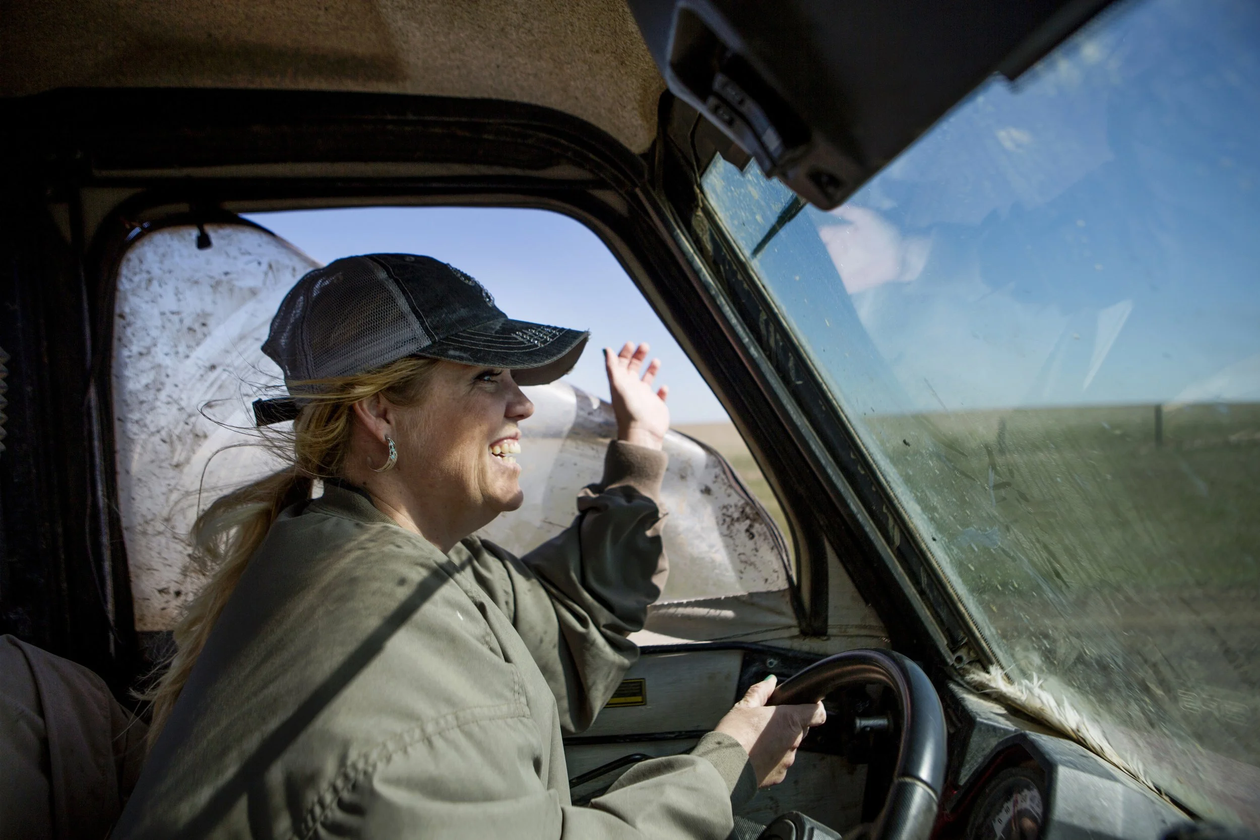 A woman with blonde hair wearing a trucker hat driving an old vehicle with a large windshield, smiling and waving at the window.
