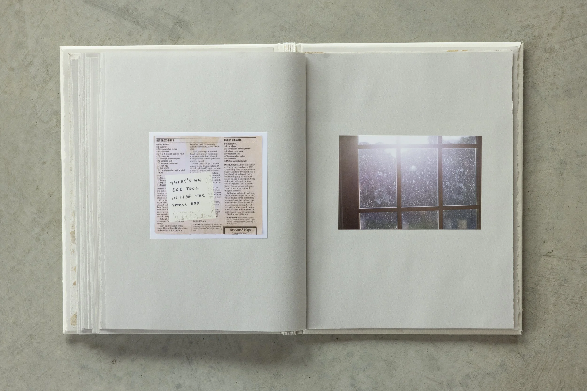 Open photo album on a gray surface, showing a recipe for 'Hot Cross Buns' and a note that says, 'Title's an egg tool inside the small box' with a list of ingredients, a photograph of a window with rain outside, and the album's pages.