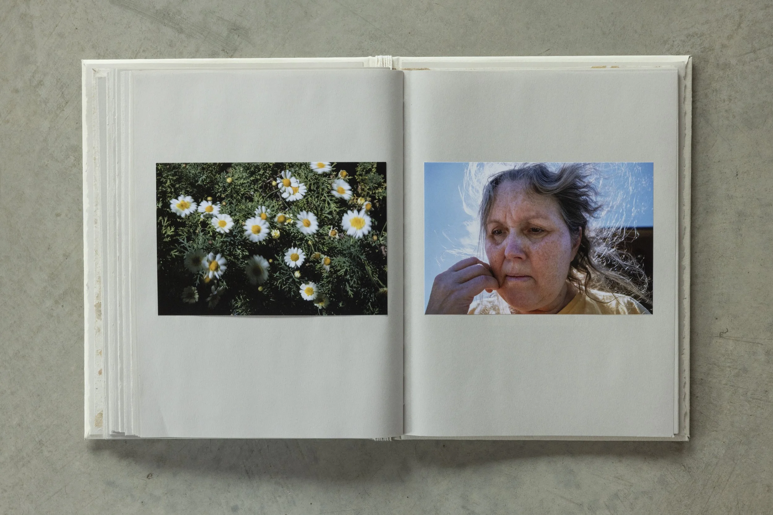 An open photo album with a picture of a woman with gray hair, and a picture of daisies with yellow centers on a dark background, placed on a gray surface.