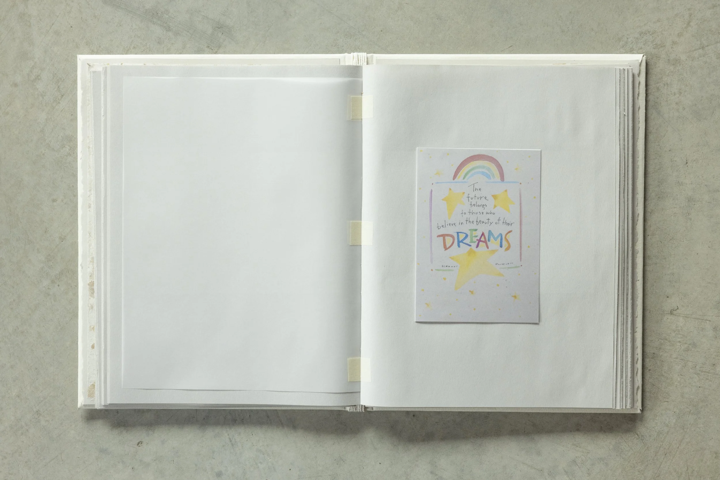 Open white scrapbook lying on a concrete surface, featuring a hand-drawn rainbow, stars, and an inspirational quote about dreams.