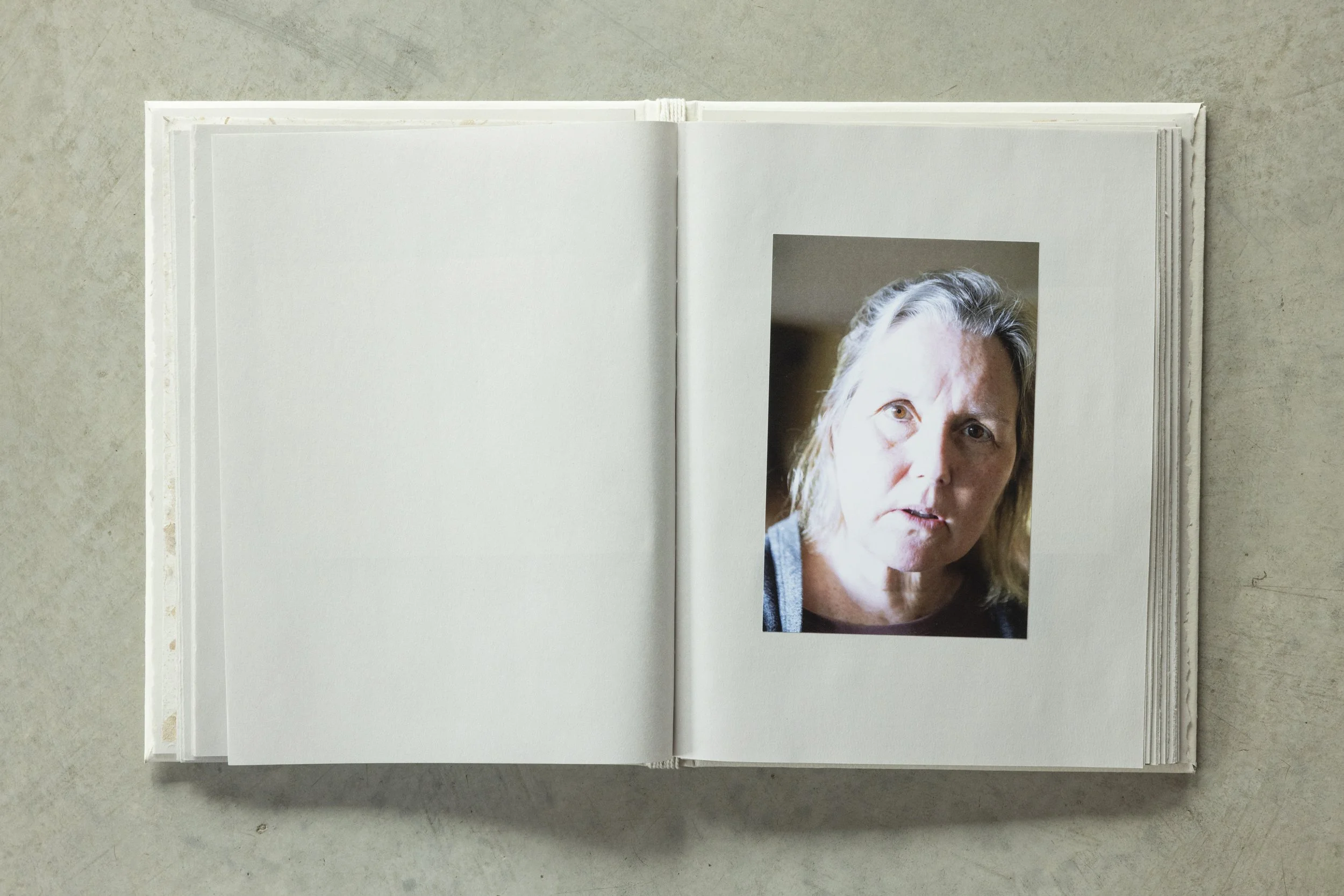 An open photo album with a portrait of a woman with short blonde hair, wearing a grey shirt, on a light grey surface.
