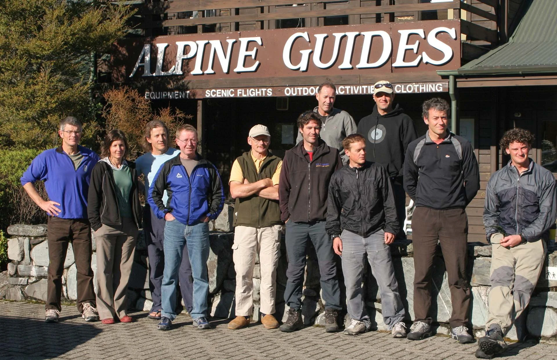 Group of people standing in front of an 'Alpine Guides' sign, wearing outdoor clothing, with a stone wall and wooden building in the background.