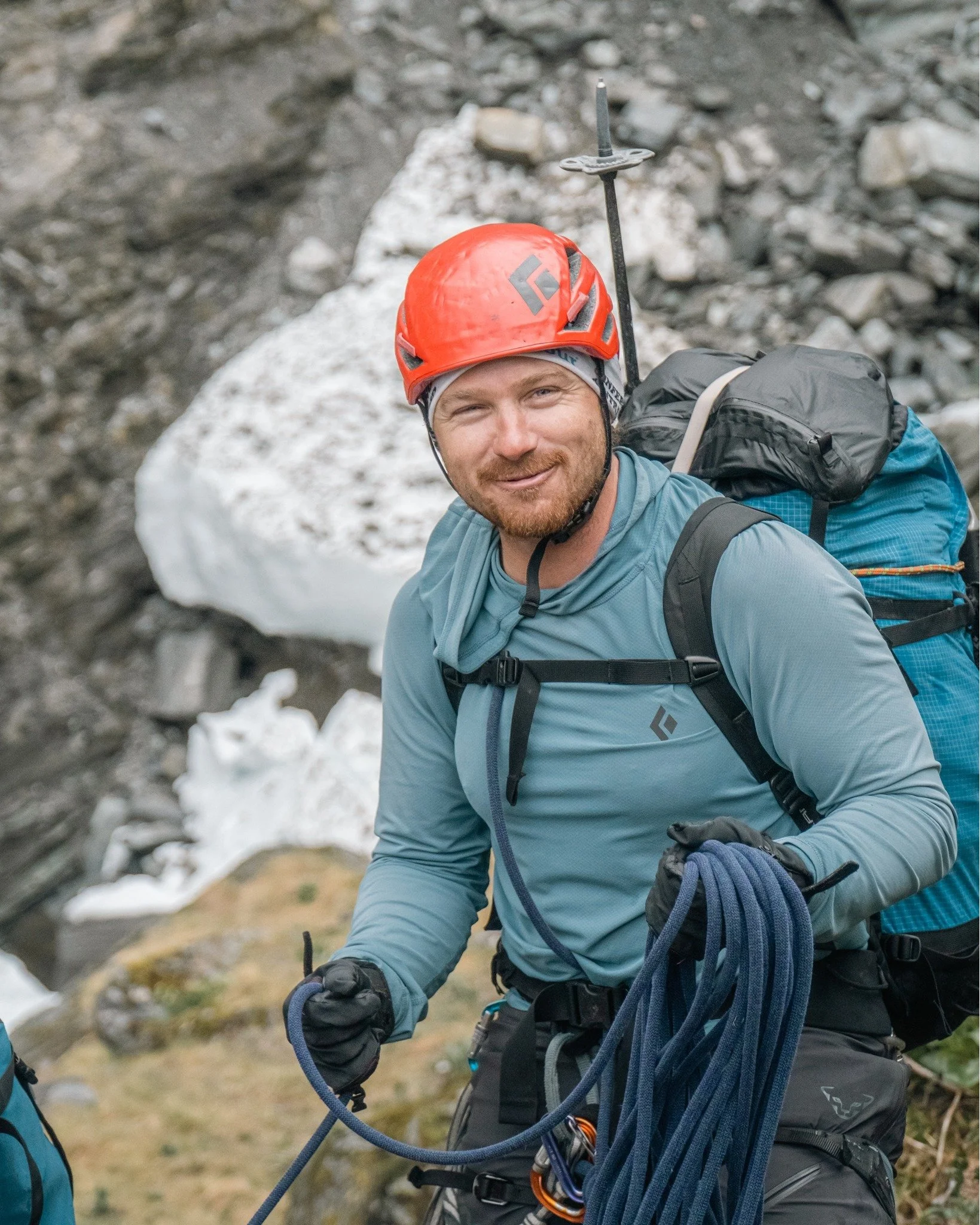 Mountain climber wearing a red helmet and teal long-sleeve shirt, holding a climbing rope, with a backpack in a rocky, snowy terrain.