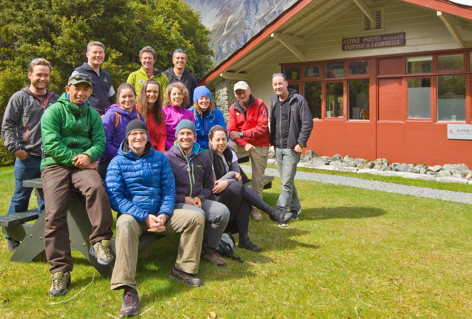 A group of people posing outdoors in front of a building labeled 'Alpine Guides'. They are dressed in outdoor gear and smiling, standing on grass with trees and mountains in the background.