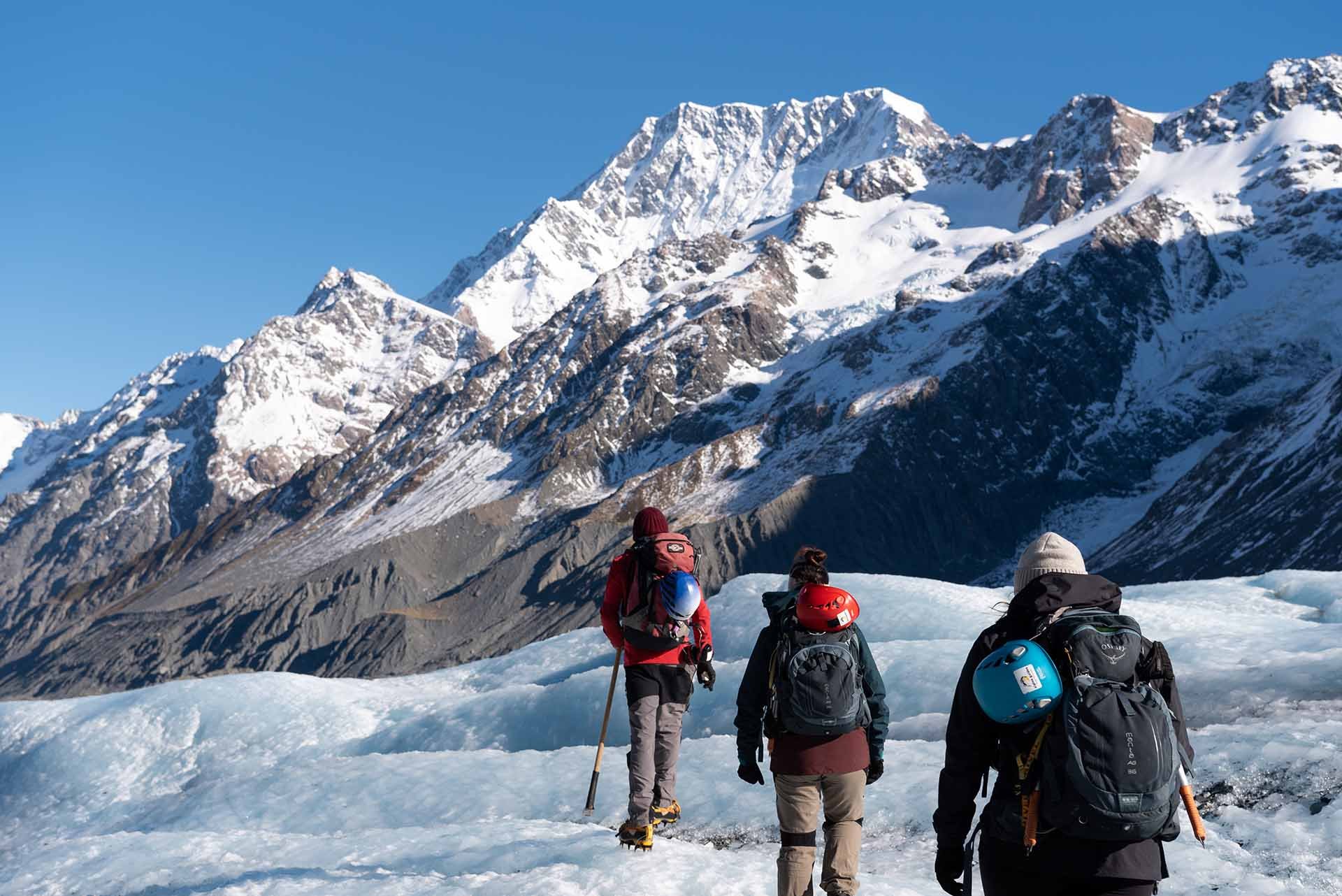 Three hikers wearing backpacks and helmets walking on a snowy glacier with snow-covered mountains in the background under a clear blue sky.