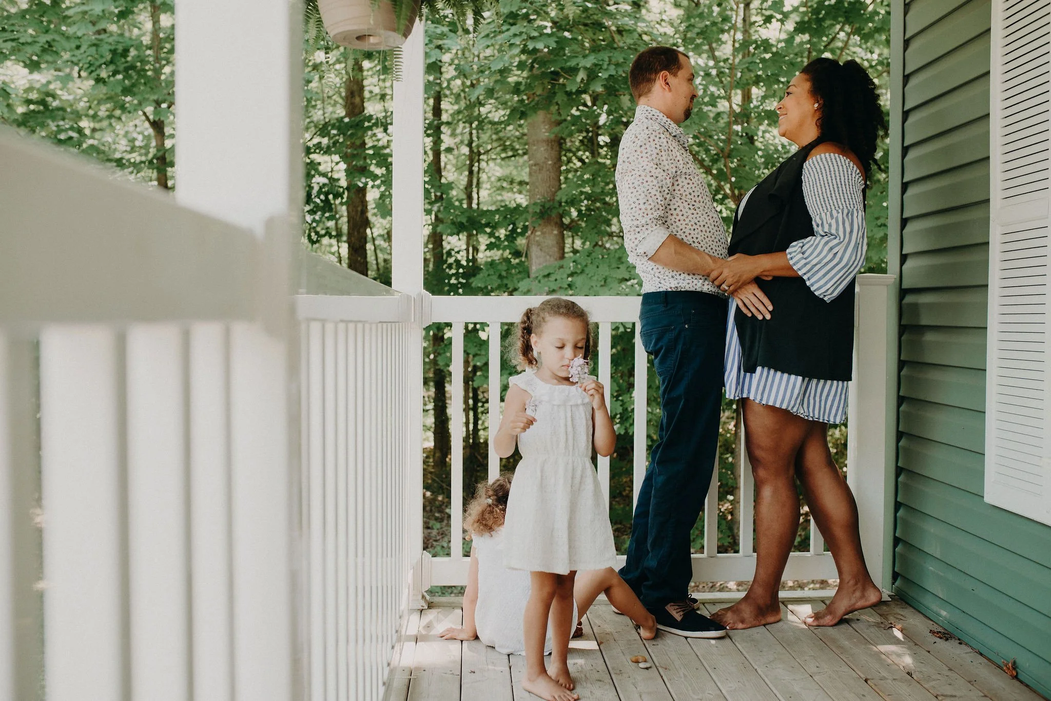 An expecting couple holds hands on a porch while their two young children play nearby.