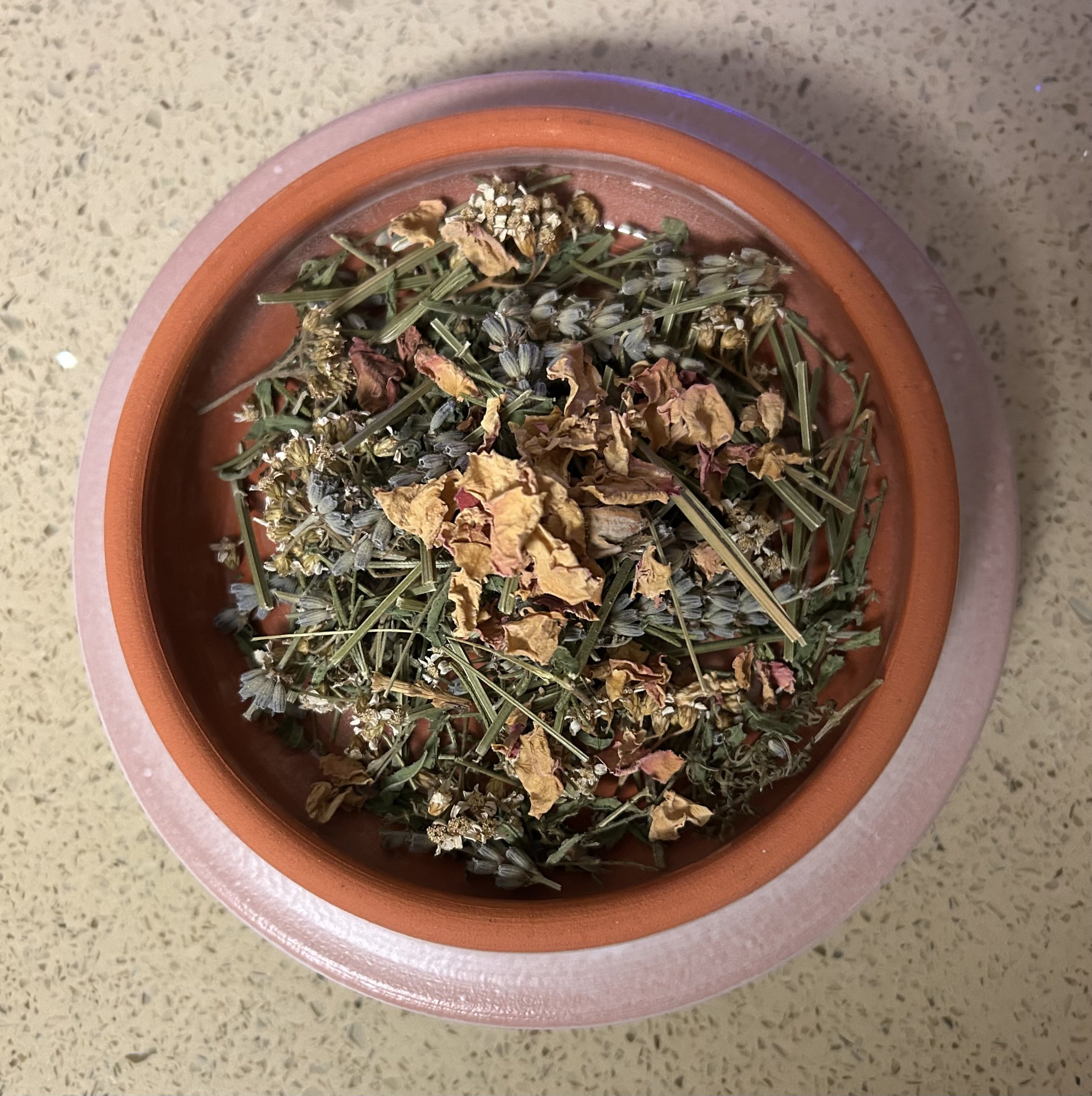 A terracotta bowl filled with dried lavender, rose petals, chamomile, and other herbs.