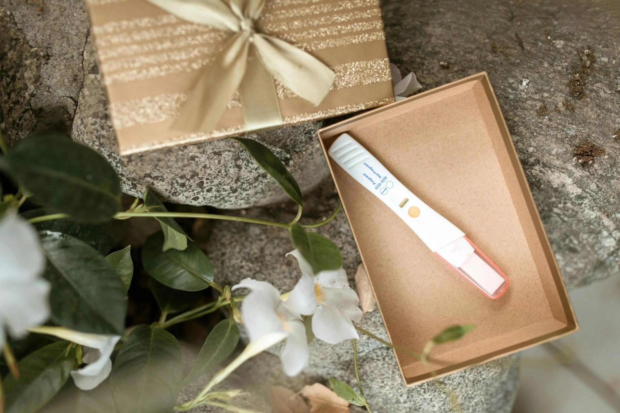 A pregnancy test resting in a small kraft box beside a gold-wrapped gift and white flowers.