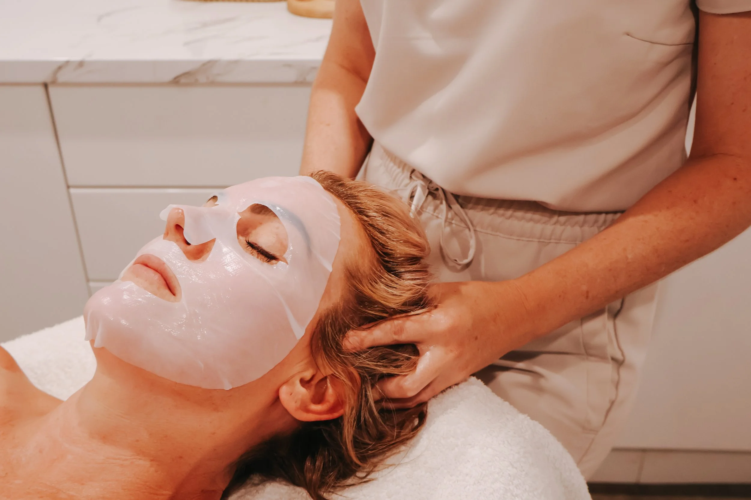 A person receiving a facial treatment with a facial mask applied, lying on a towel with a professional administering the treatment in a spa or clinic setting.