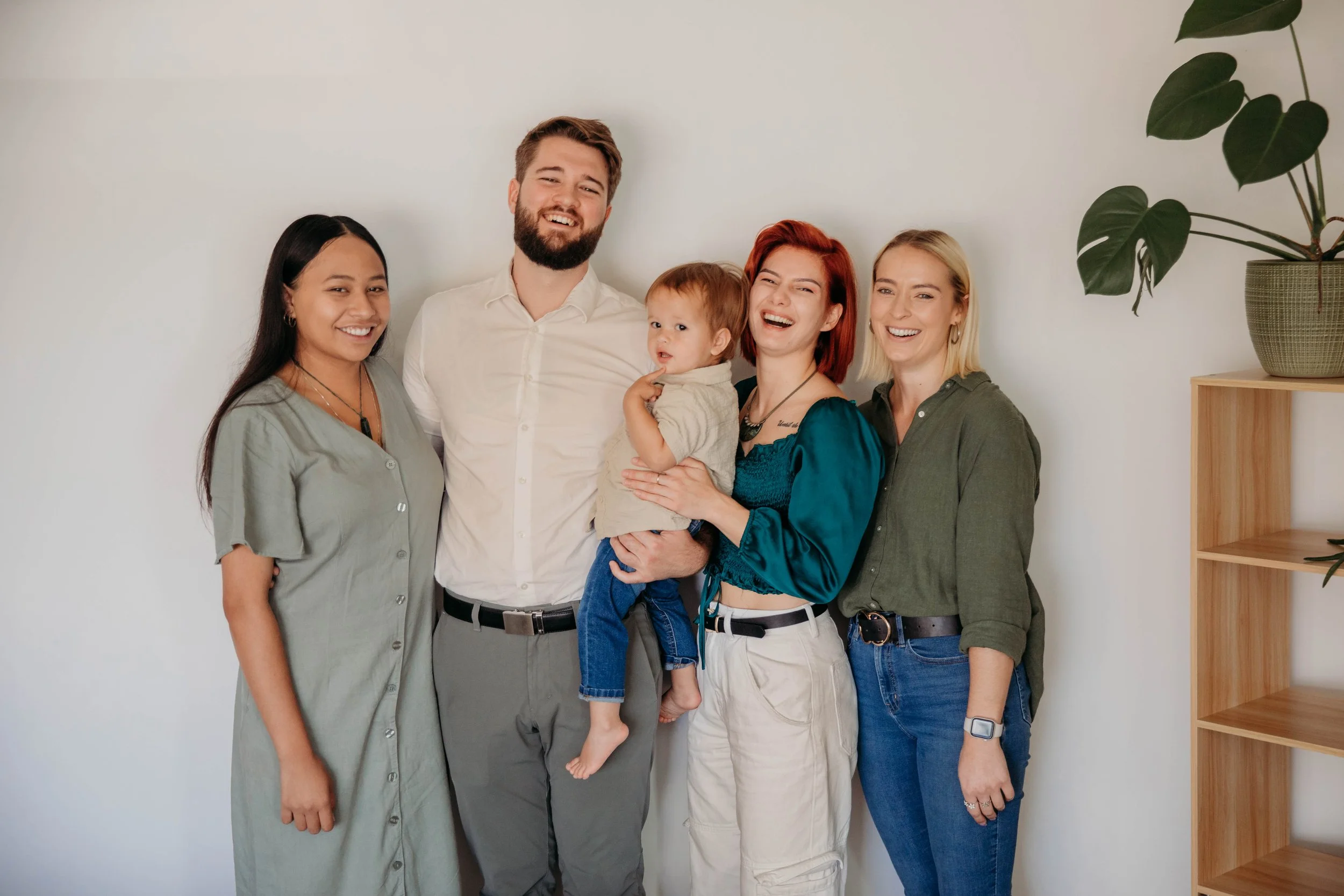 Group of five people standing together and smiling, with one person holding a young child, against a plain white wall with wooden shelving and a plant on the right.
