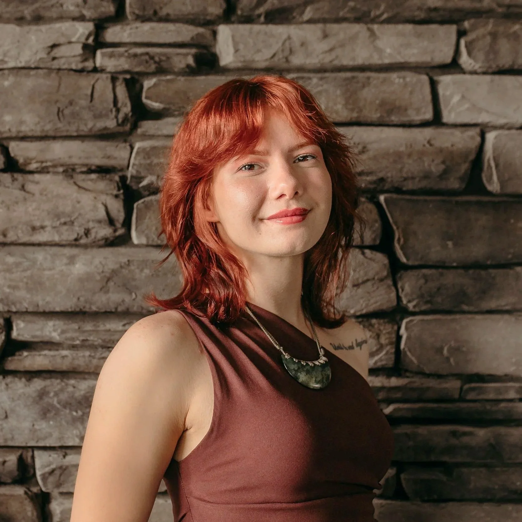 A young woman with short, red hair smiling slightly, wearing a teal blouse with ruffled neckline and a large green stone necklace, standing against a plain white wall with a partial view of a green plant leaf on the right.