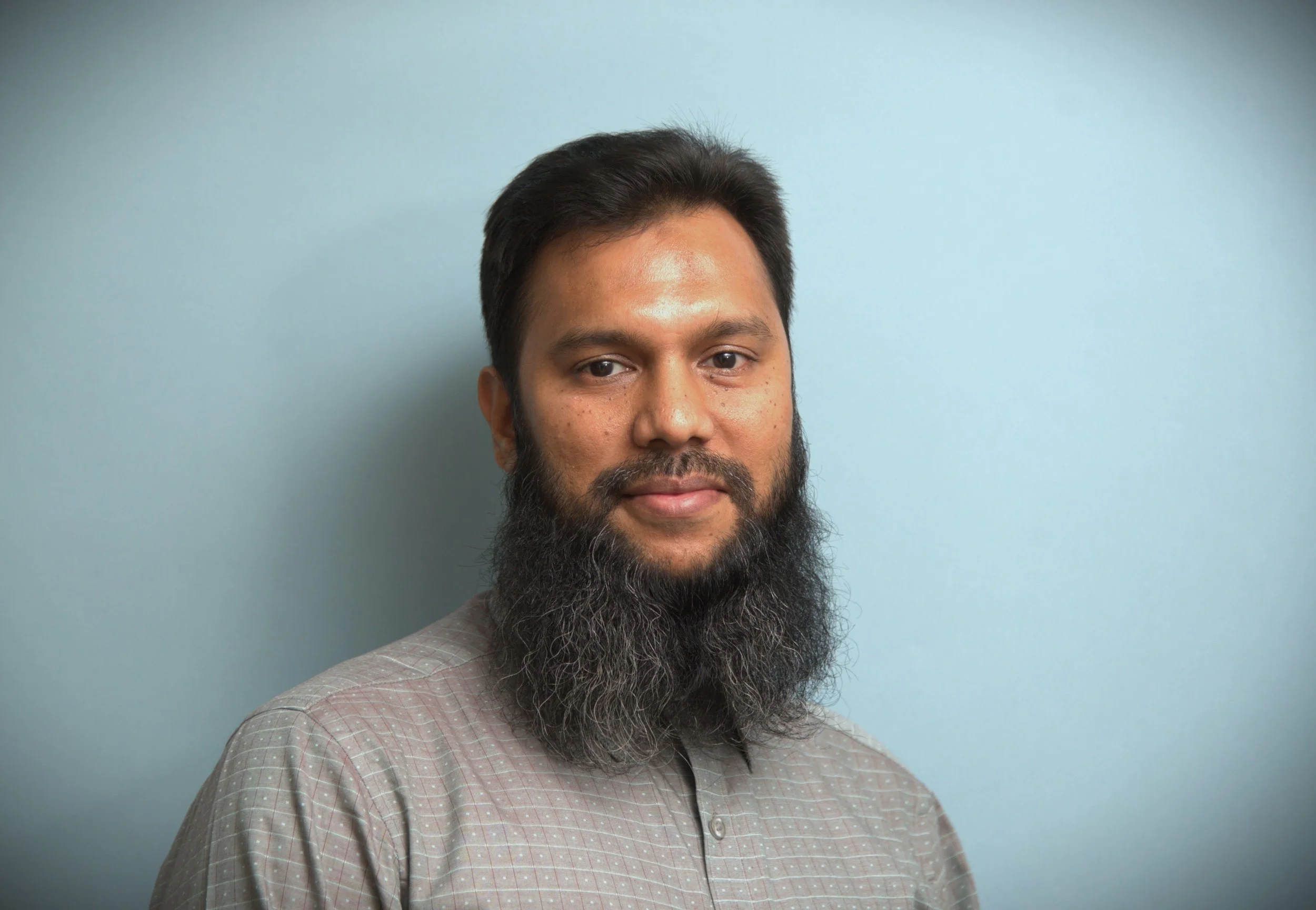 Man med skägg och randig skjorta mot ljusblå bakgrund.