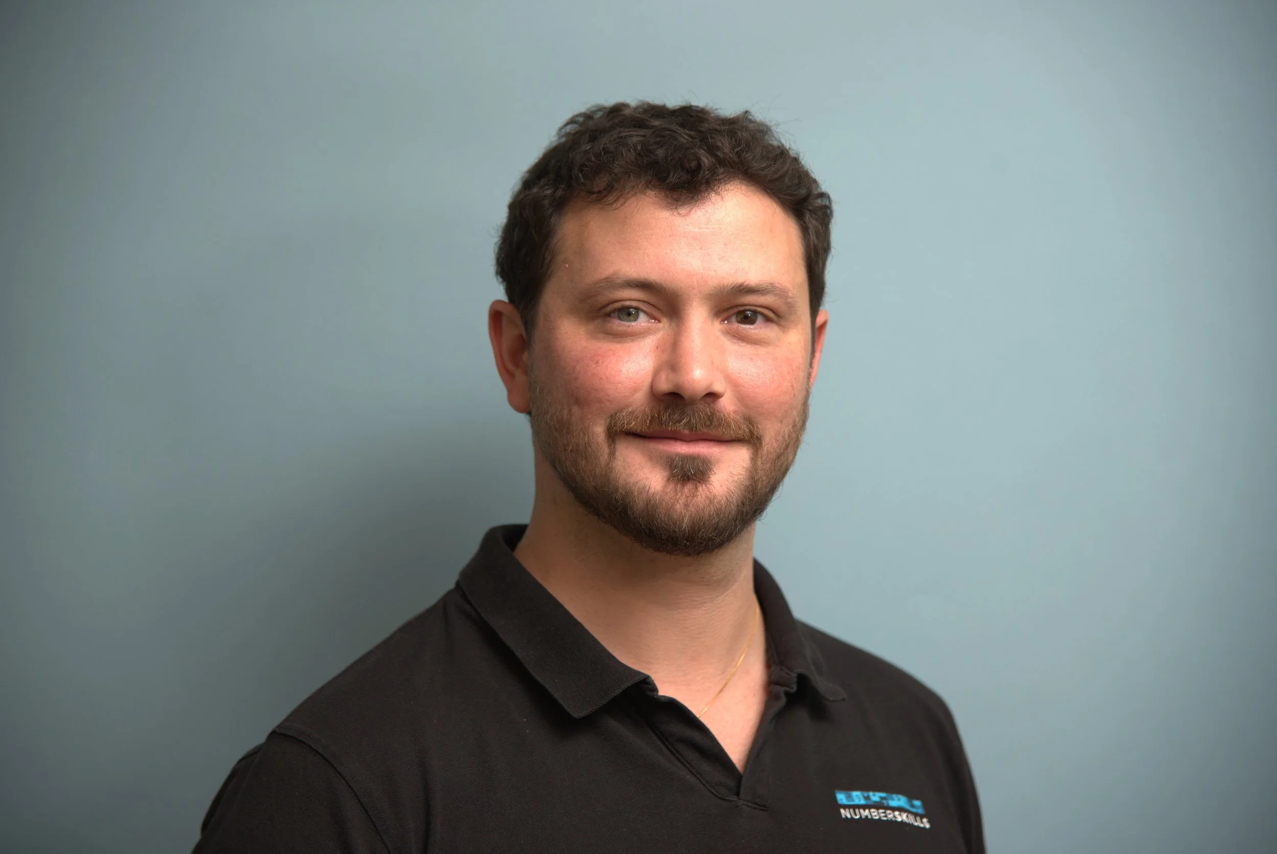 Porträtt av en man med mörkt hår och skägg, klädd i en ljusgrå polotröja, mot en ljusblå bakgrund.