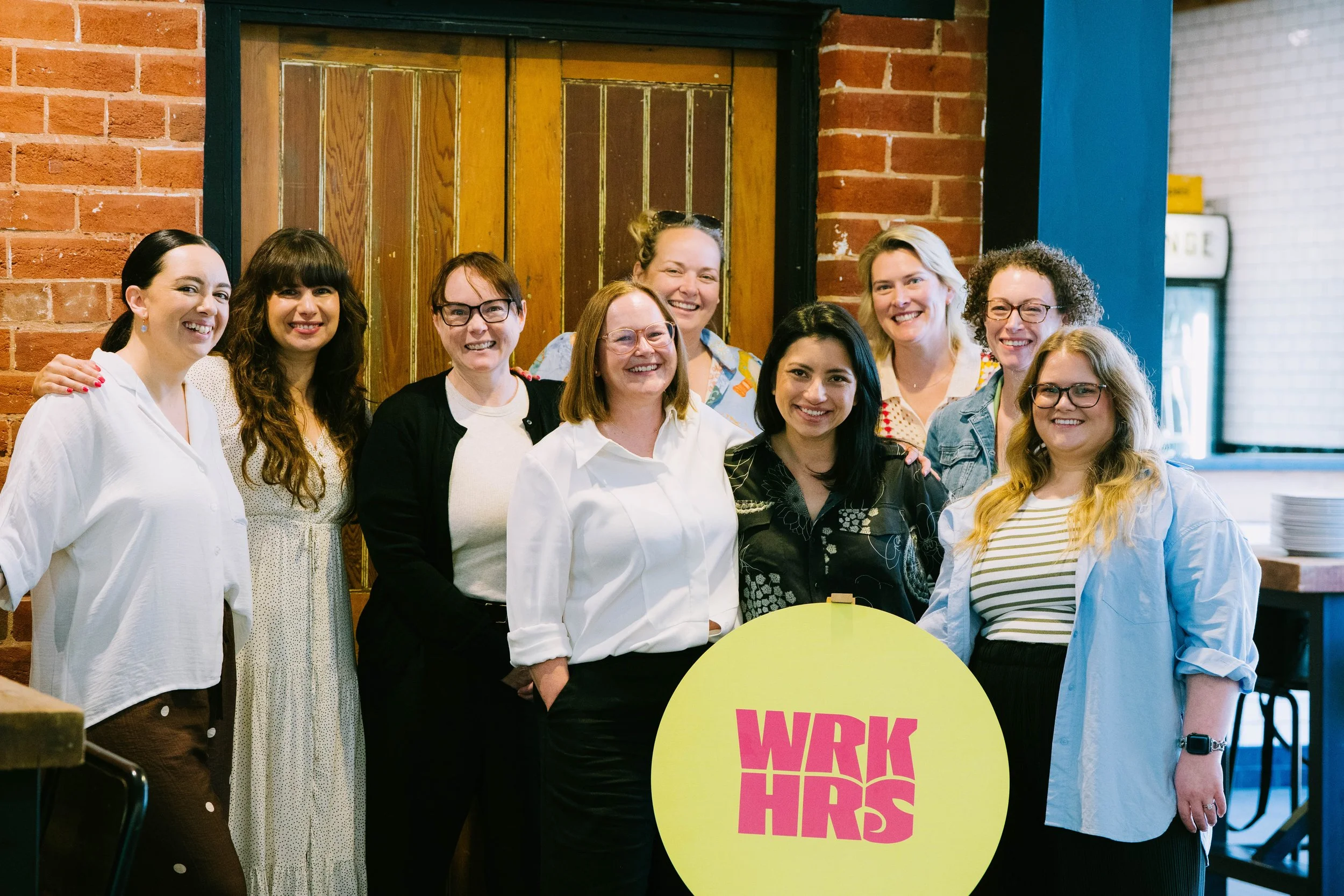 A group of eleven women standing together in a room with brick walls, smiling at the camera. They are dressed in casual to business-casual clothing. One of them holds a yellow circular sign with pink text that reads "WRK HRS."