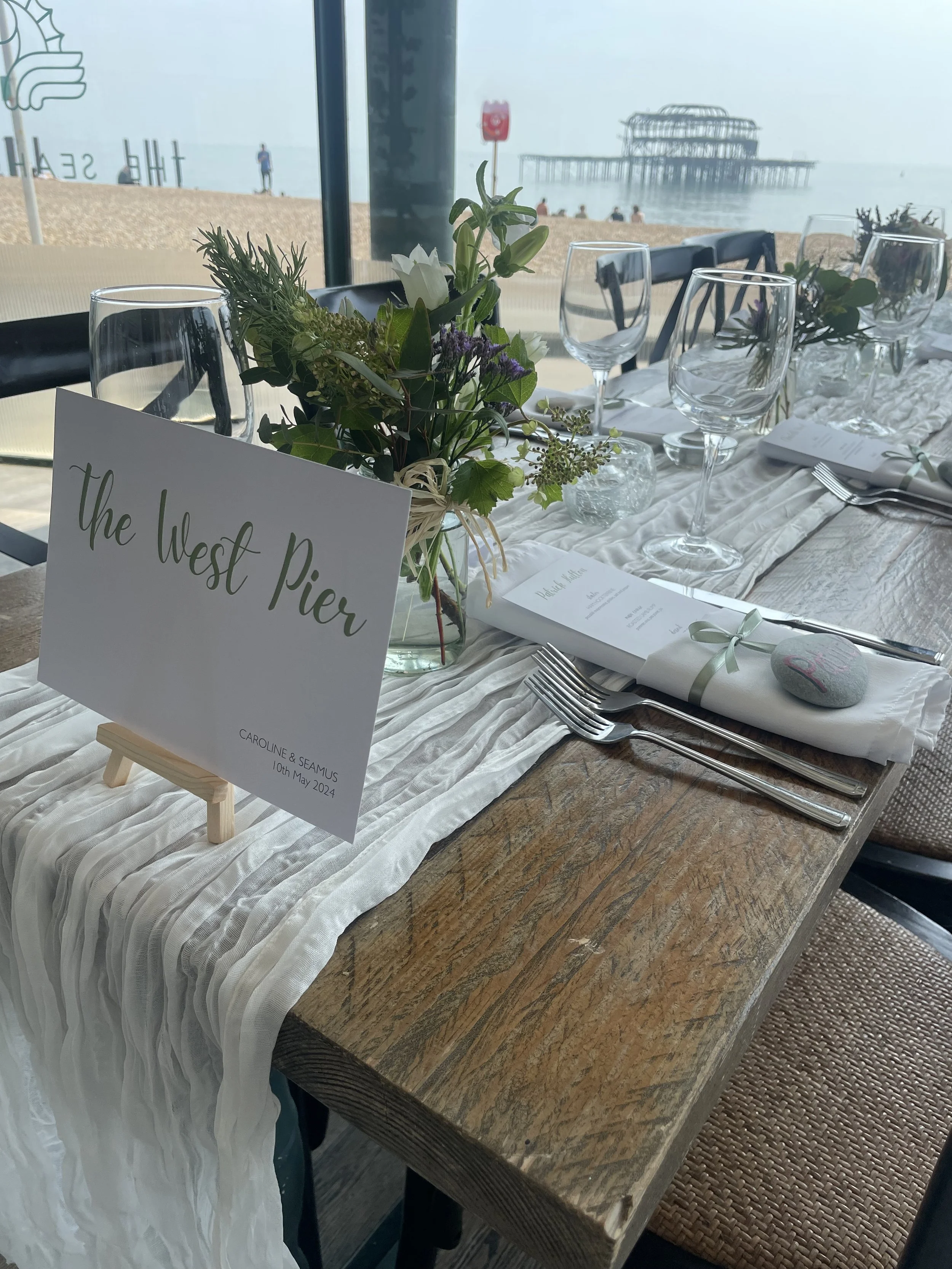 Wedding table setup by the beach with a sign reading "the West Pier," decorated with flowers, wine glasses, and neatly folded napkins, overlooking the pier and ocean.