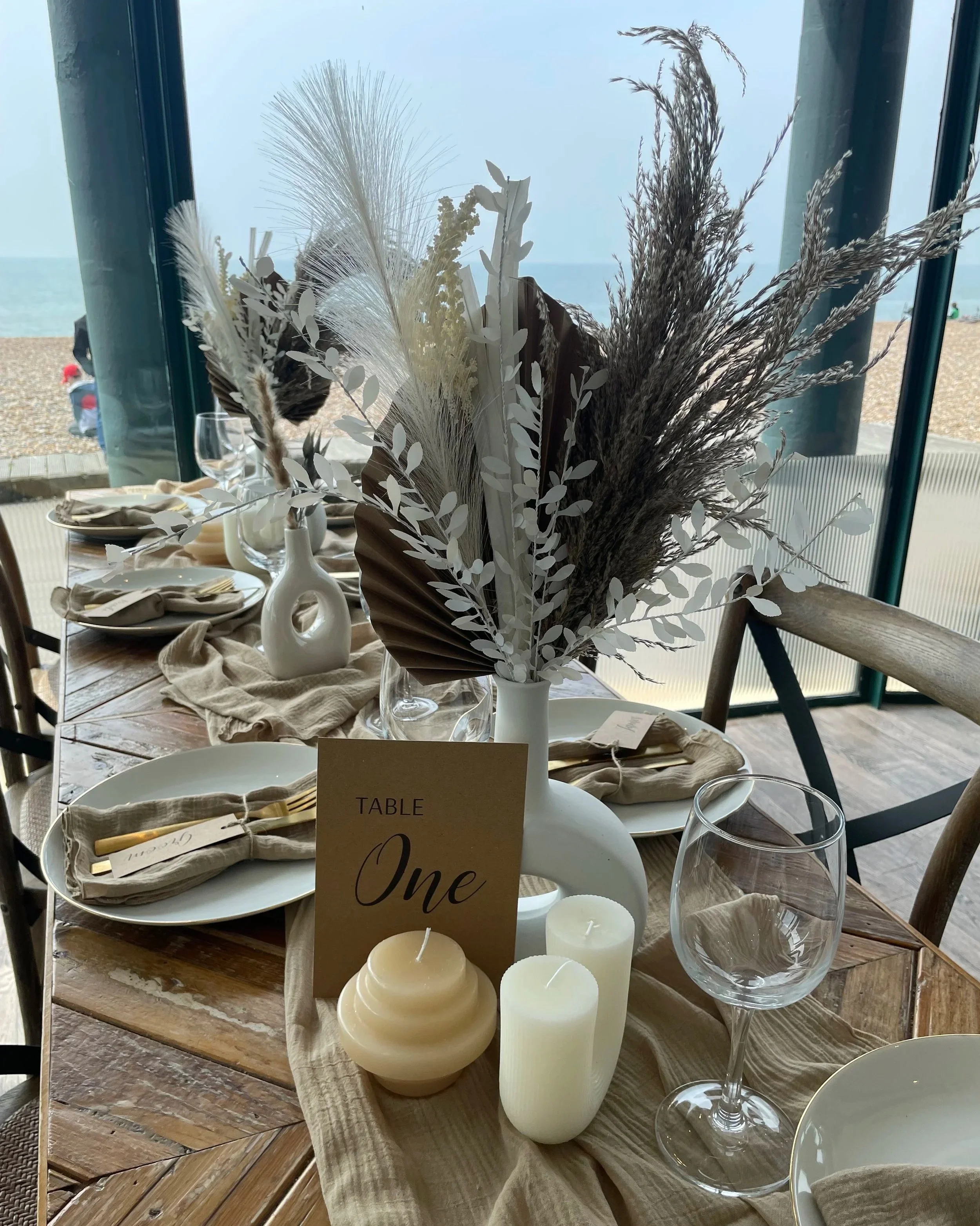 Table setting with a white vase holding dried ornamental grasses and leaves, candles, wine glass, and beige napkins with place cards, set near a window with a view of a beach and ocean.
