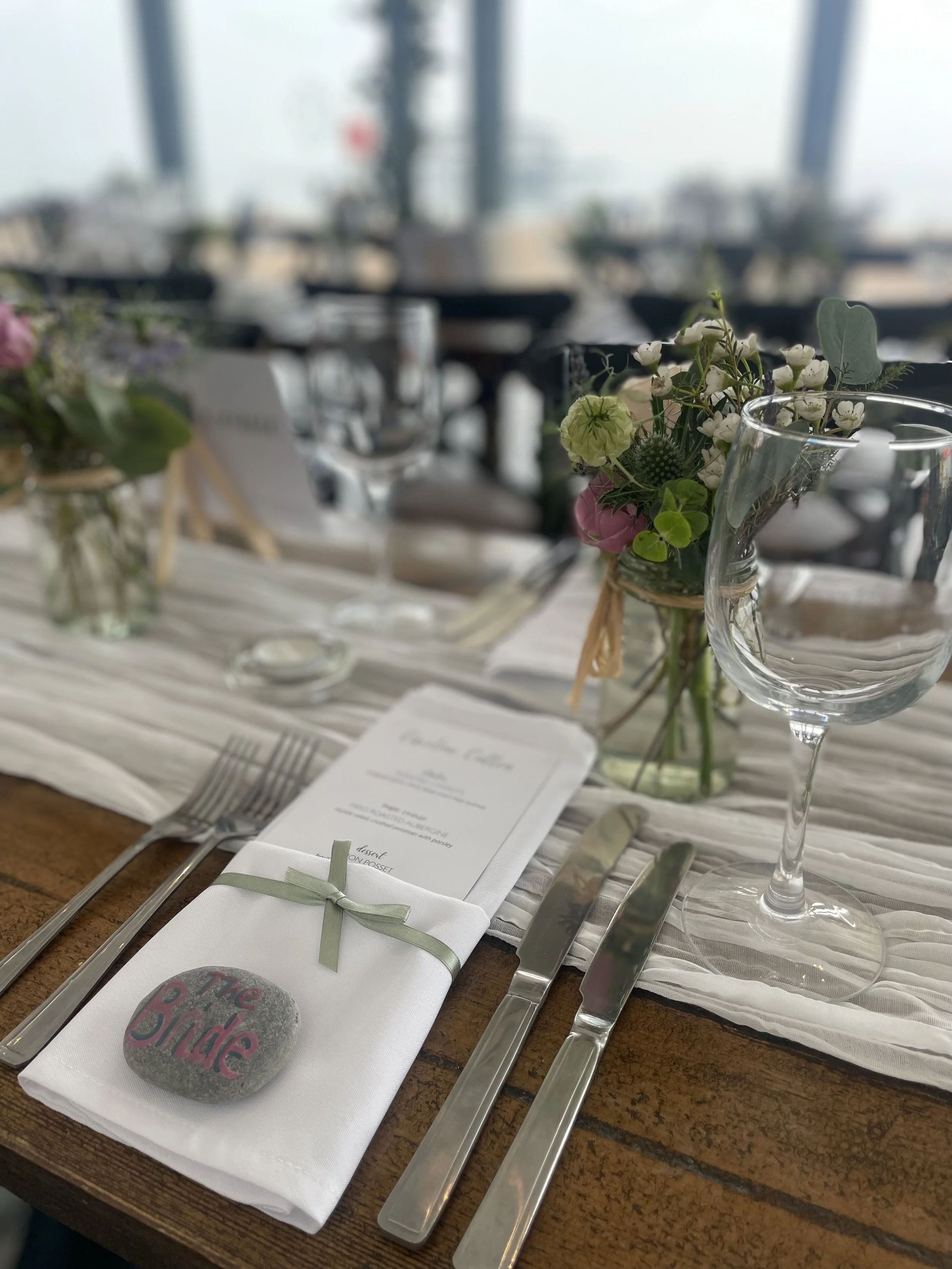 A decorated table set for a wedding, with a white napkin tied with a green ribbon and a painted rock with "The Bride" written on it, surrounded by flowers in glass jars, a wine glass, and cutlery.