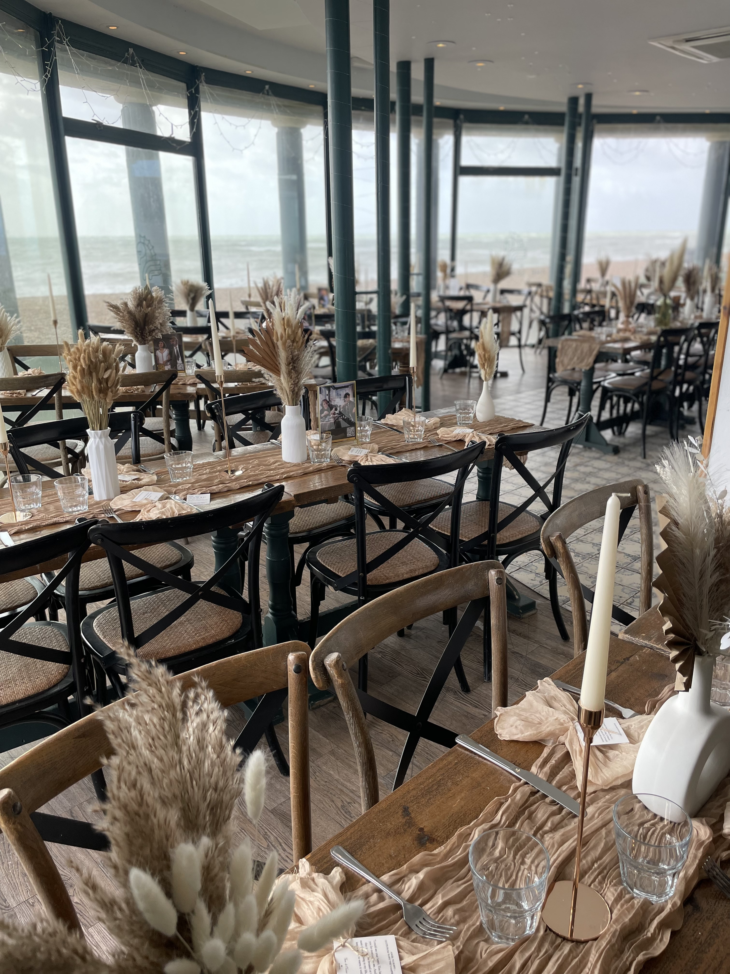 Decorated seaside restaurant with tables set for dining, featuring vases with dried pampas grass and candles, large windows overlooking the ocean, daytime setting.