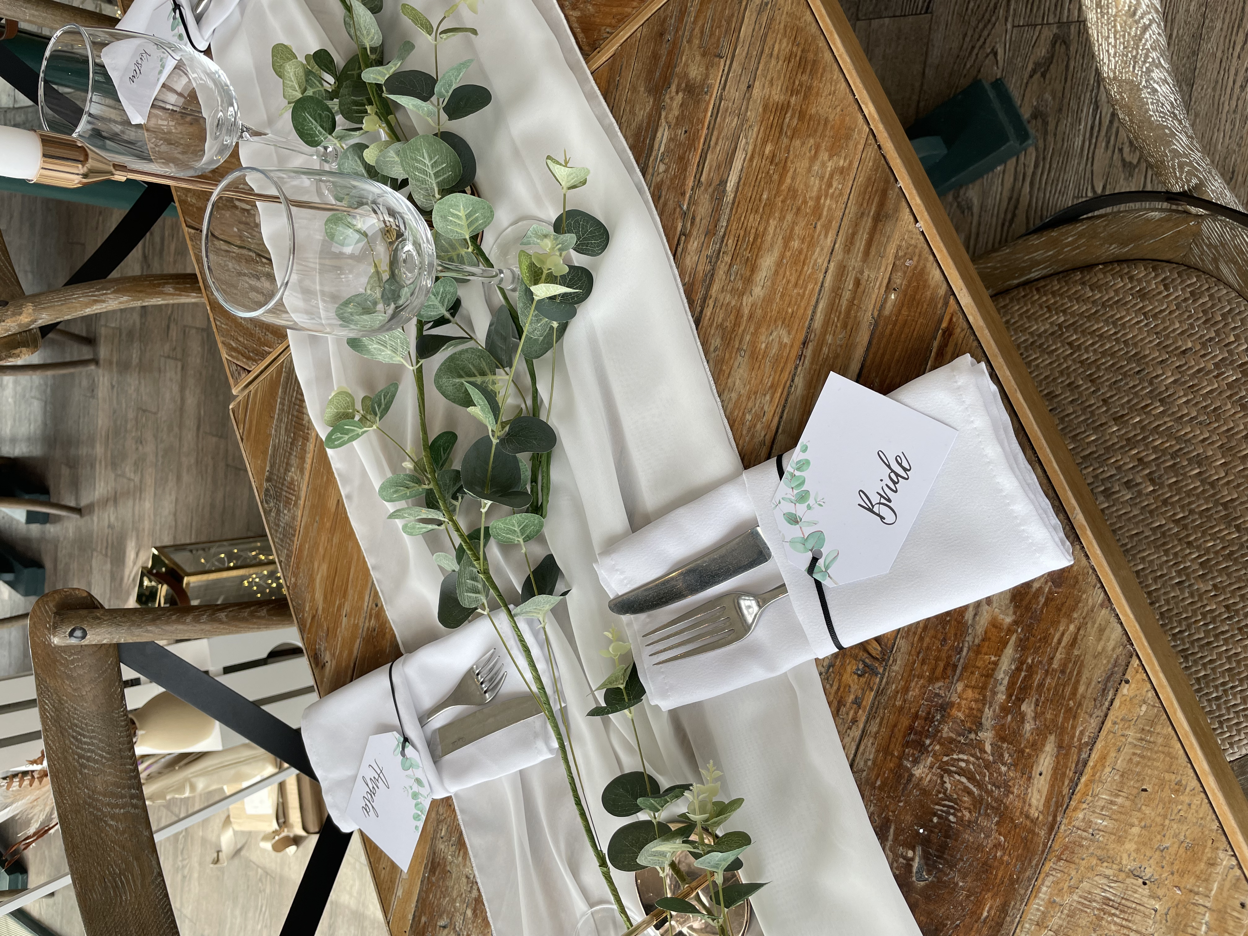 A wedding table setting with a white table runner, greenery decoration, place setting with a white napkin, fork and knife, and a table card labeled 'Bride'.