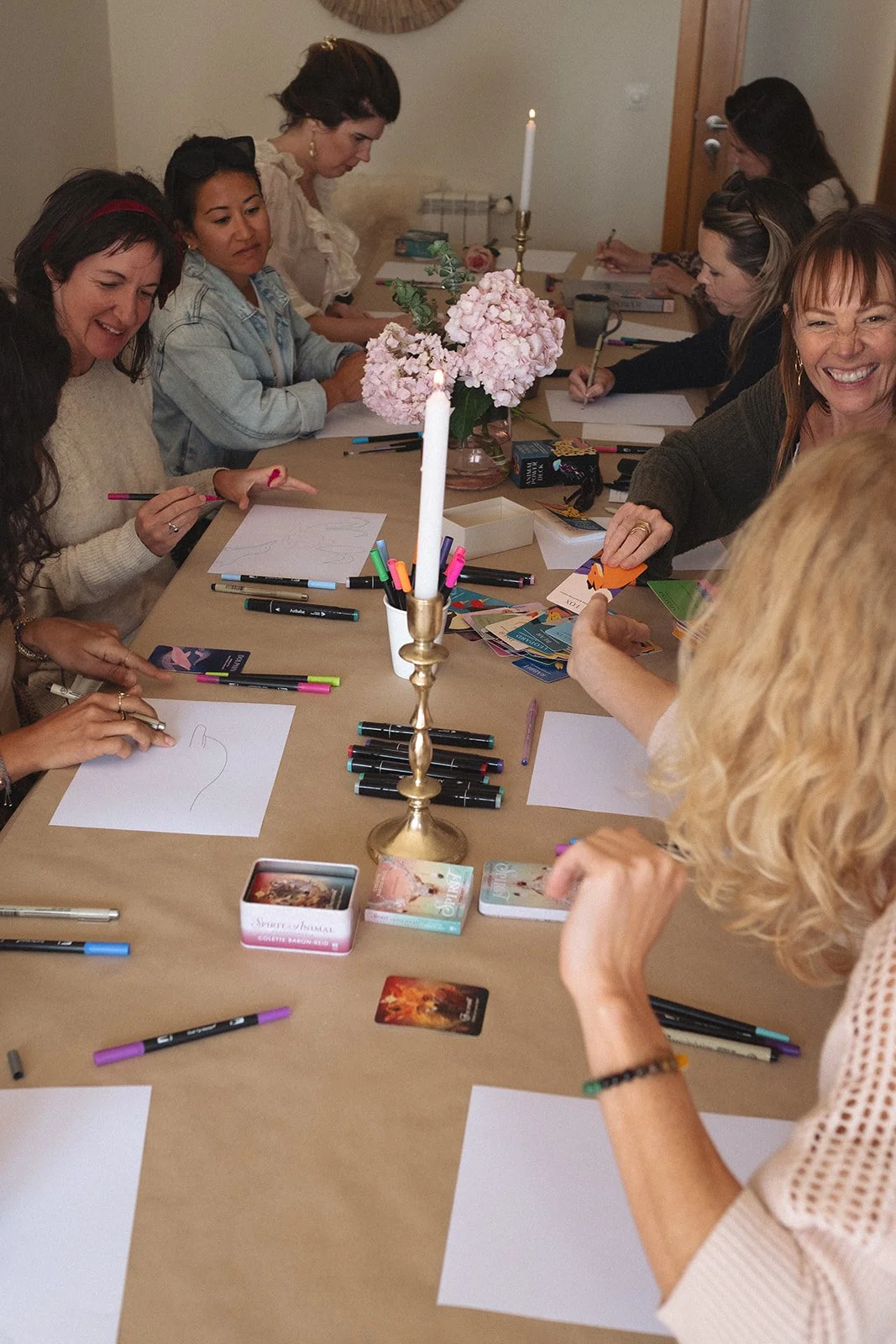 A group of women sitting around a table, engaged in a creative or card reading activity, with pens, blank sheets of paper, cards, a candle, and a pink flower centerpiece on the table.
