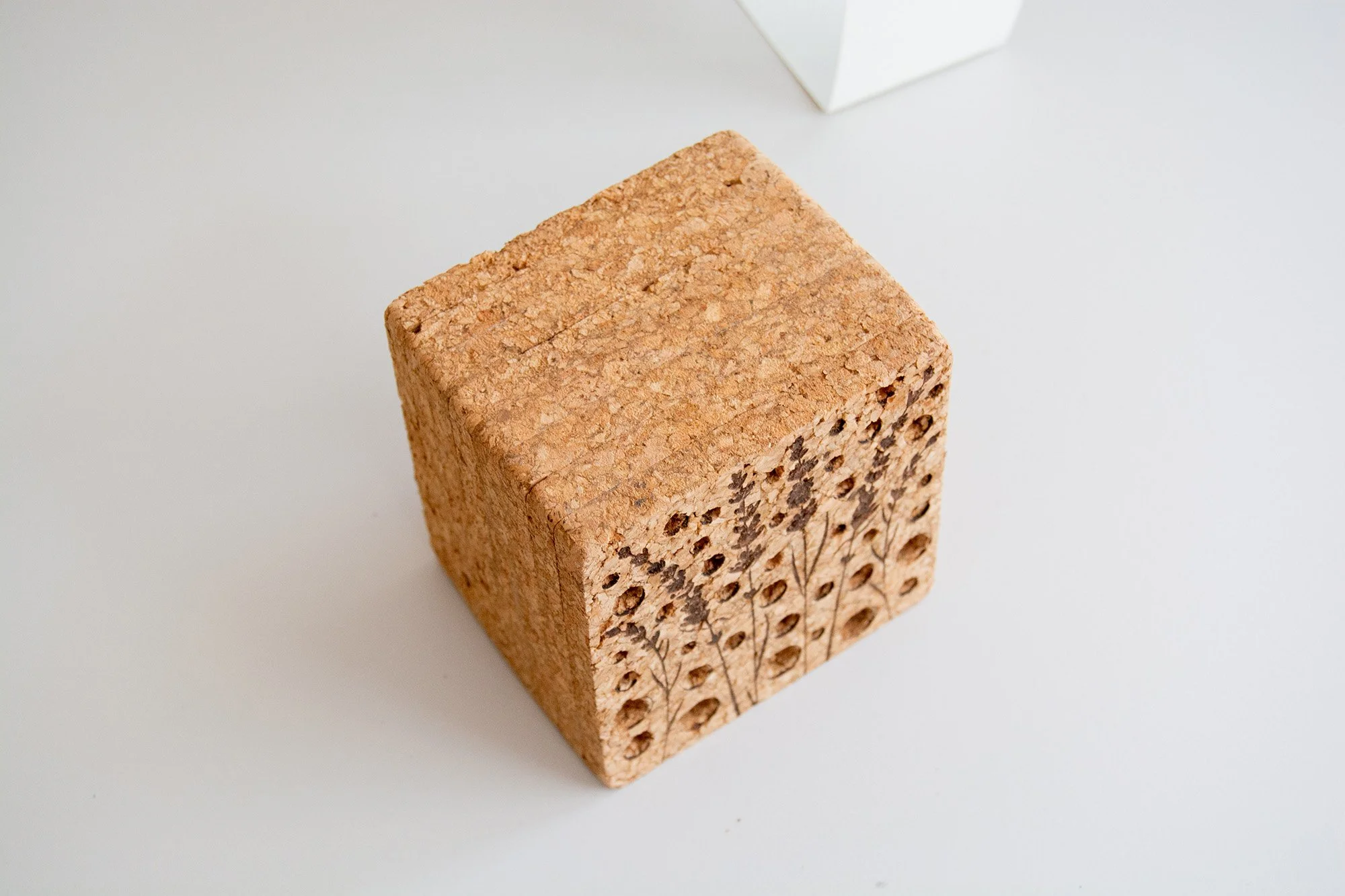 Caja de madera con forma de casa y tubos de diferentes tamaños, rodeada de paja y sobre fondo neutro. Detrás una caja de cartón con una etiqueta con patrón.