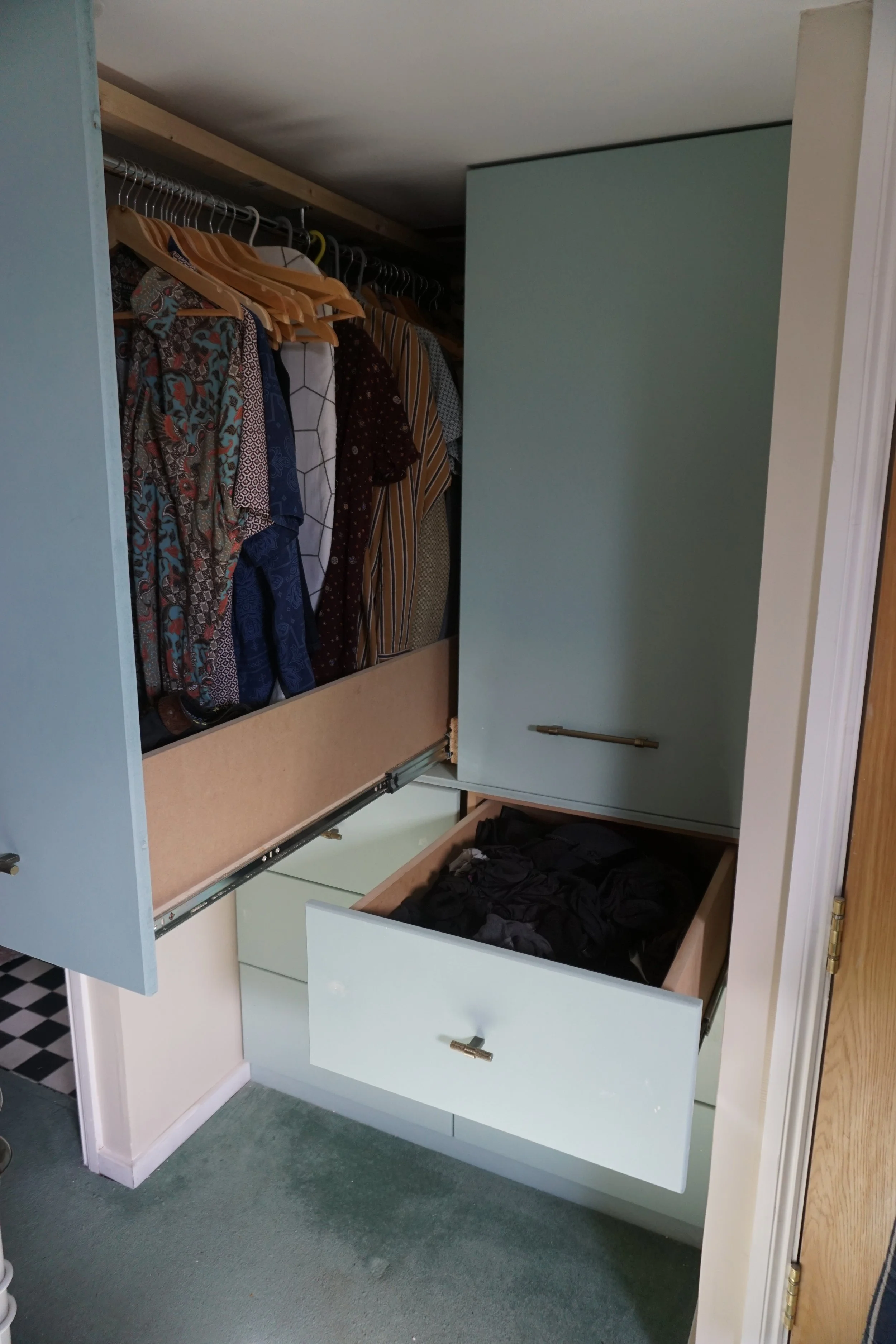 Open blue wardrobe with hanging shirts and a drawer with black clothes inside, set against a green carpeted floor.