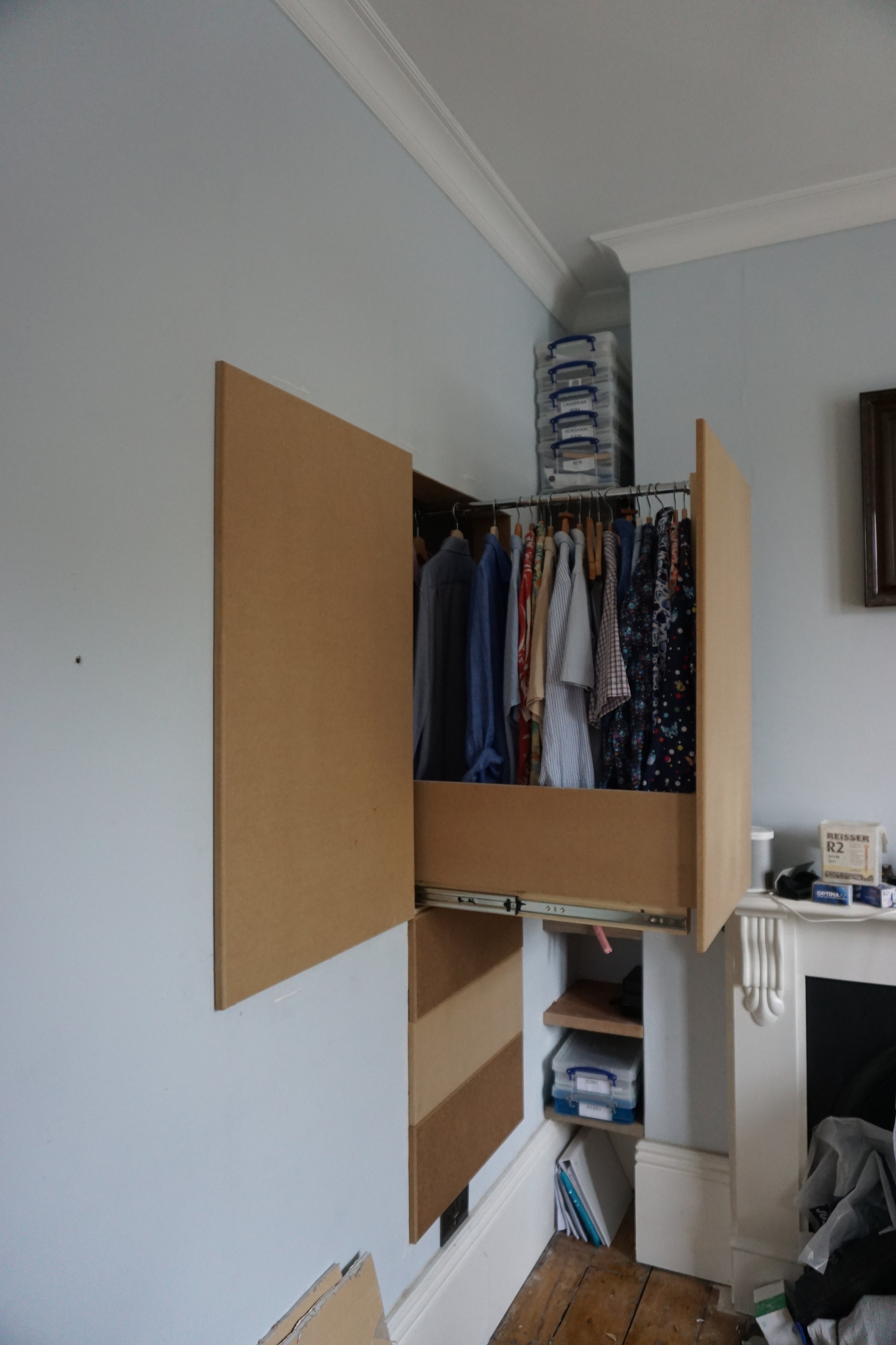 Open closet with hanging shirts and folded items, above which are plastic storage drawers, in a room with light blue walls and wooden floor.
