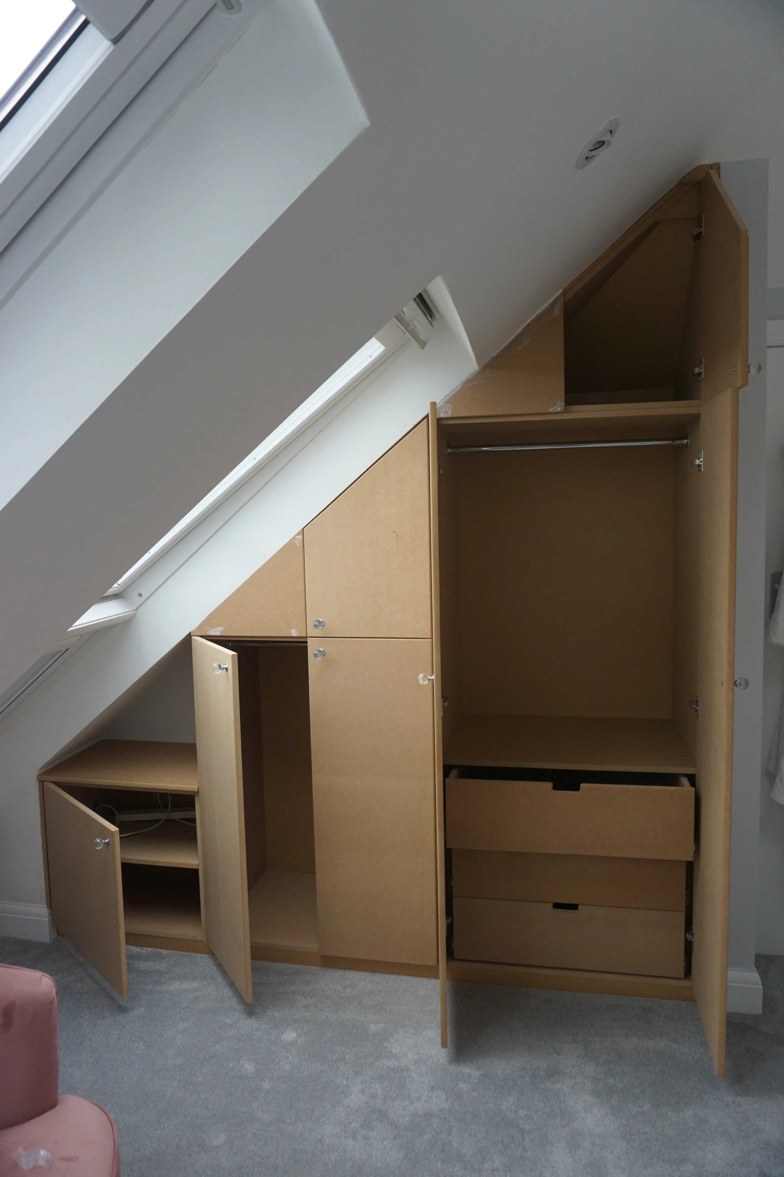 Wooden cupboards with multiple compartments and doors, located under a sloped ceiling with a window and exposed vent, on a gray carpeted floor.